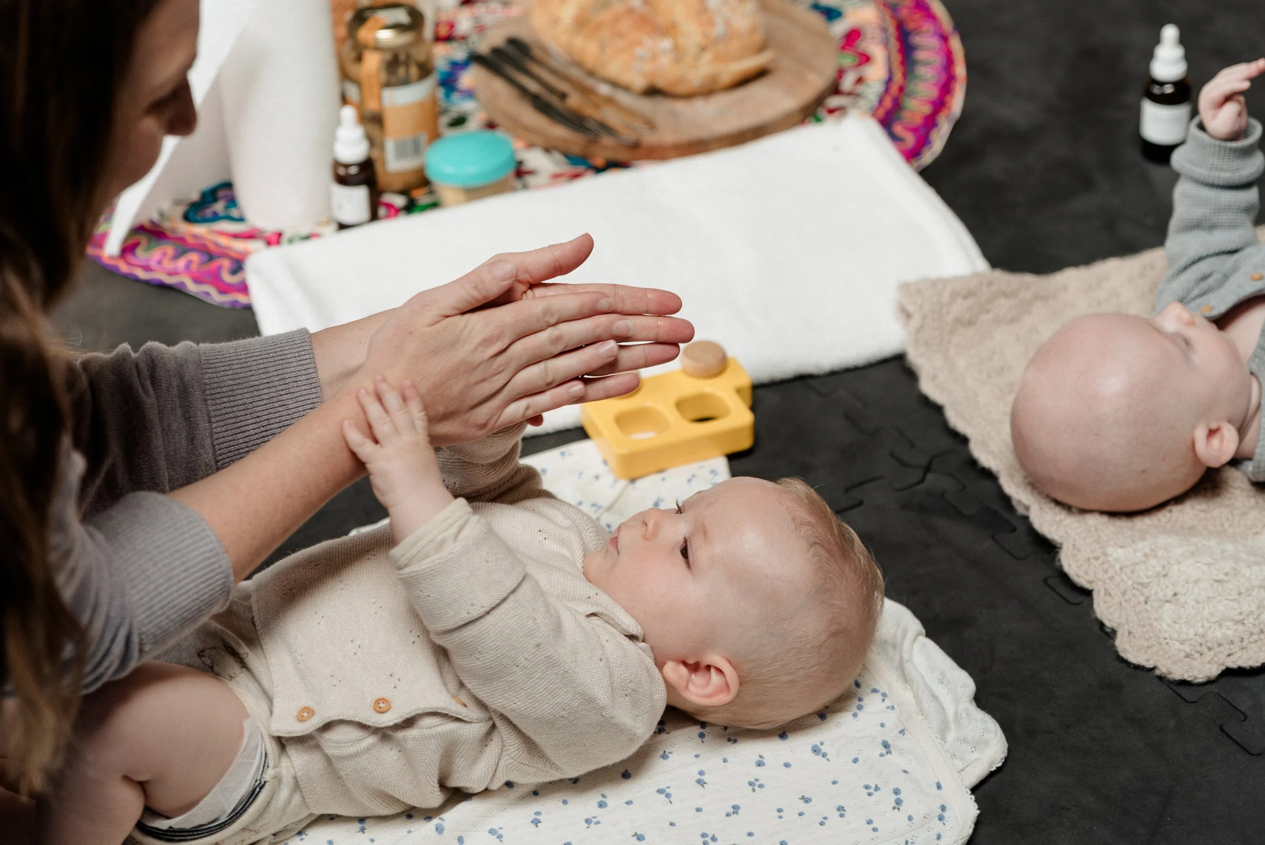 Two infants lying on their backs on white mats, with a woman clapping her hands near one baby and the other baby looking up. The background includes bottles, a colourful cloth, and a wooden plate with bread.