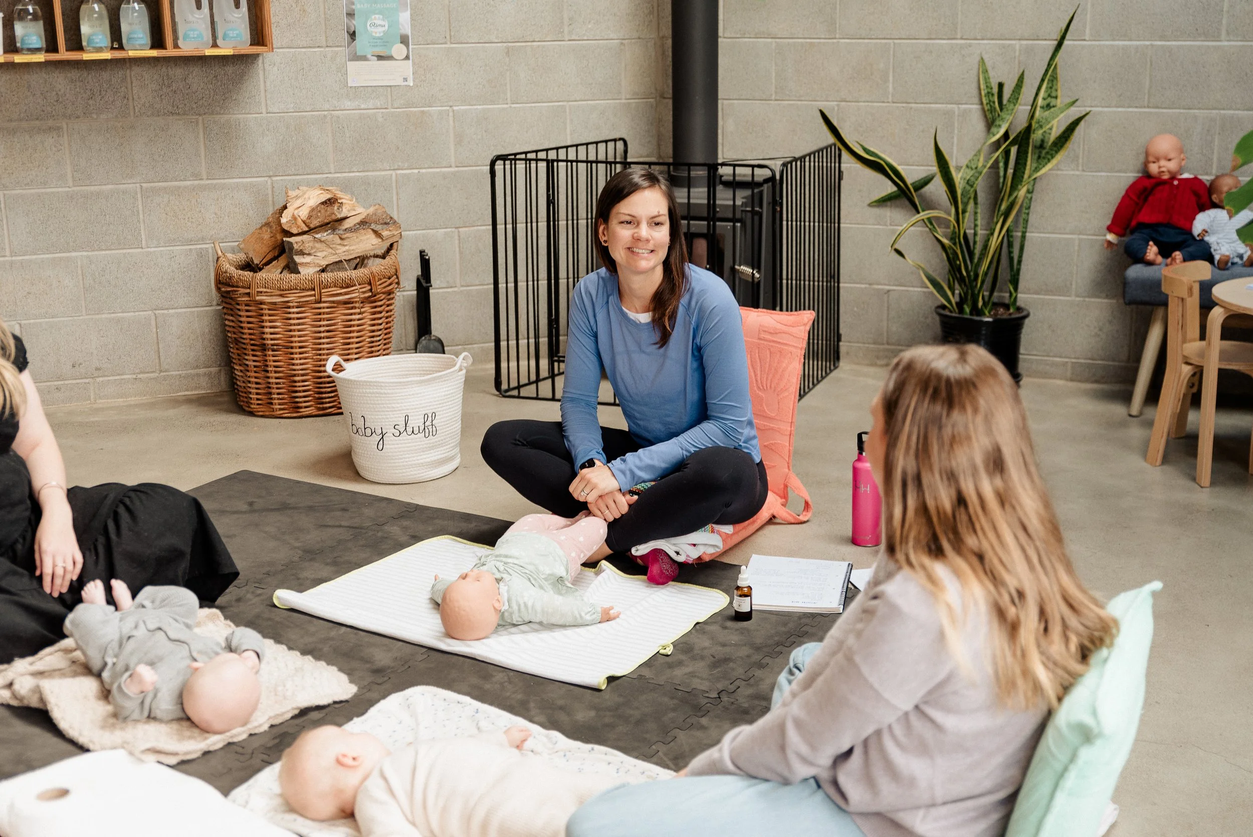 A woman with dark hair and a blue long sleeve shirt sitting cross-legged on the floor, smiling, in a room with children and babies. There are toys, a basket filled with firewood, a plant, and a doll on a chair in the background.