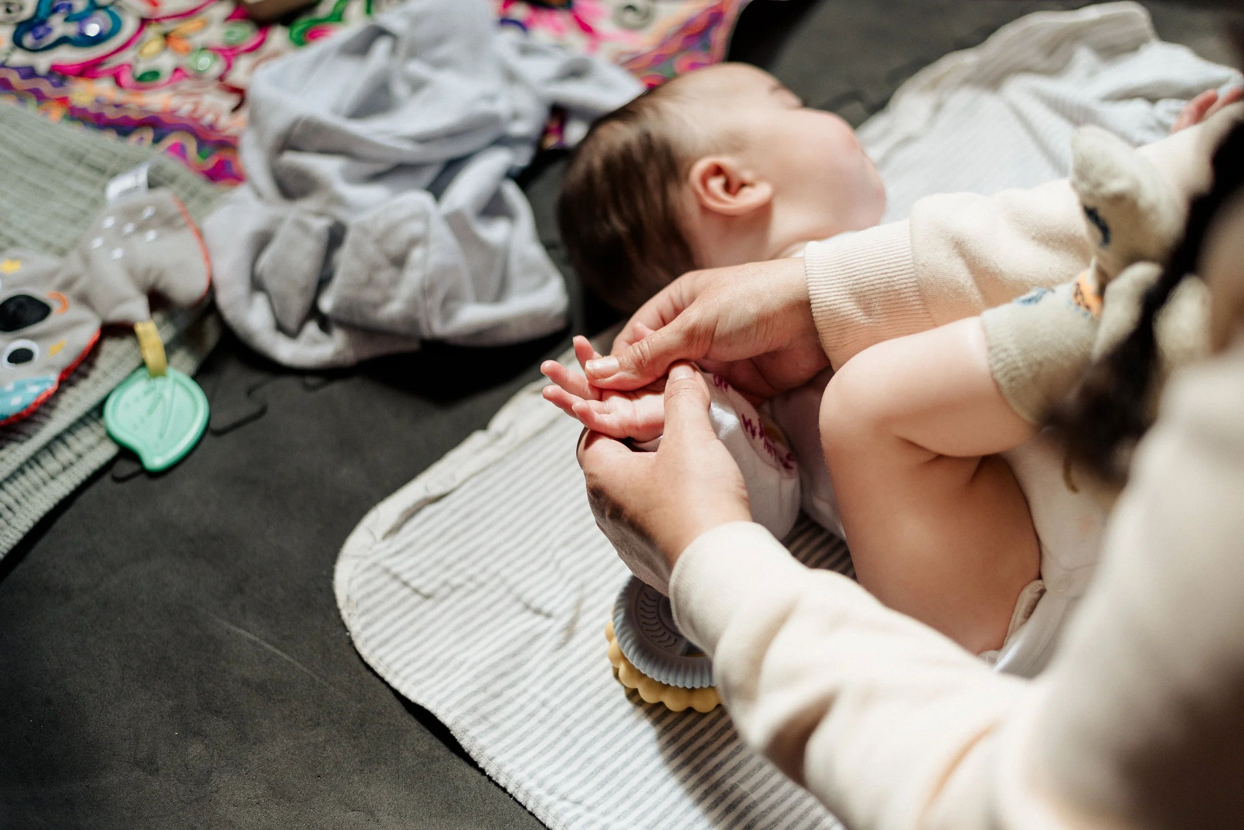A white baby lies on a striped rug on the floor, being gently held and having its hand massaged by an adult. The baby is resting peacefully looking away, surrounded by scattered blankets and toys.