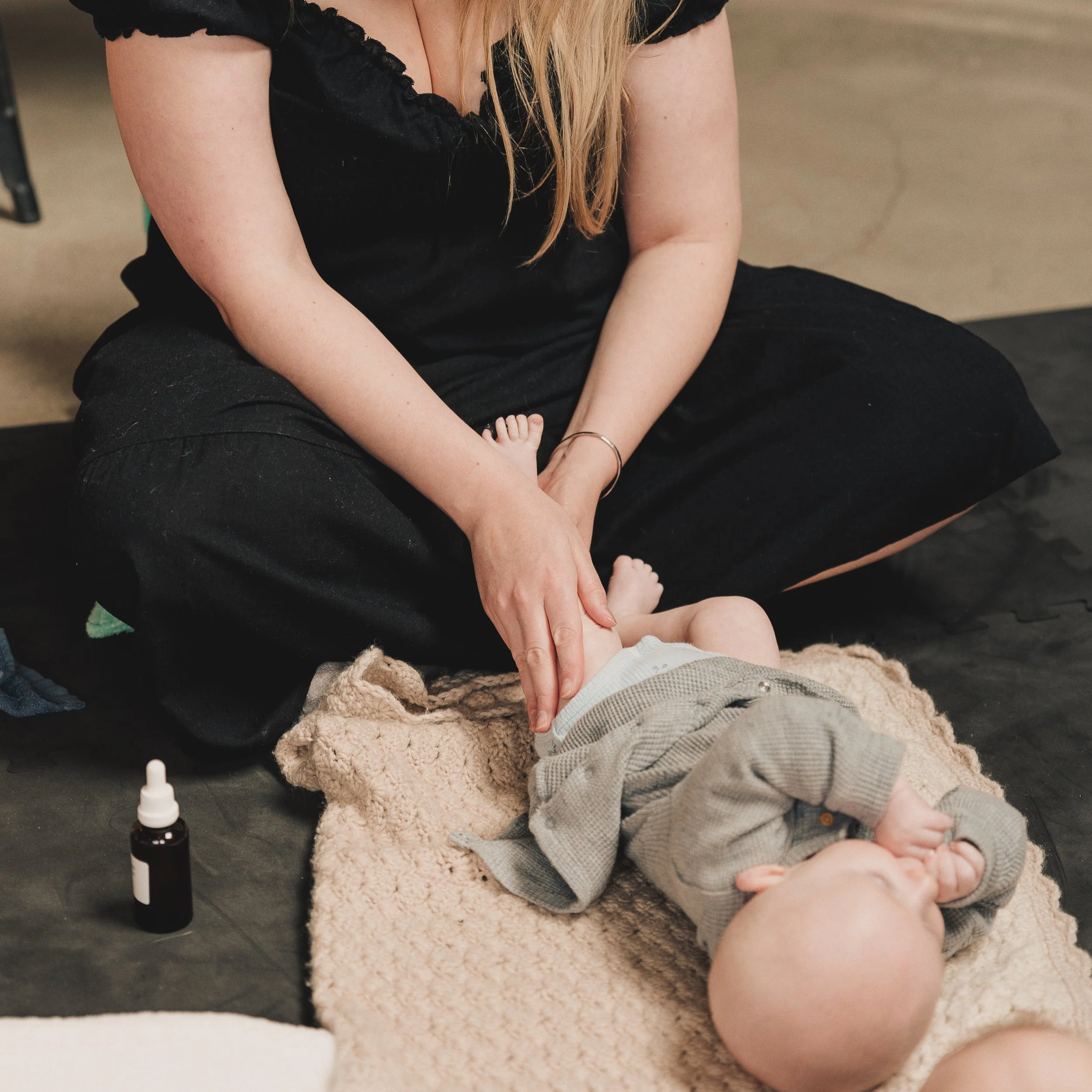 A woman in black clothing is giving a leg massage to her baby, who is lying on a beige, textured blanket. There is a small bottle containing oil with a dropper -top nearby.