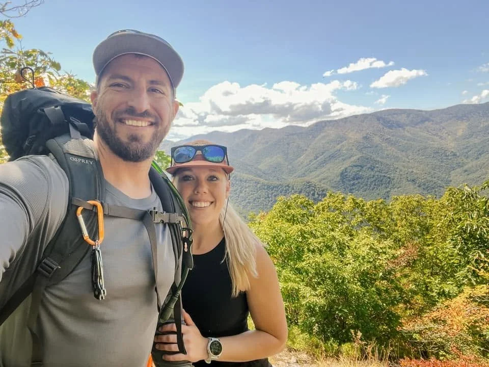A smiling man and woman hiking outdoors with a mountain range and partly cloudy sky in the background.