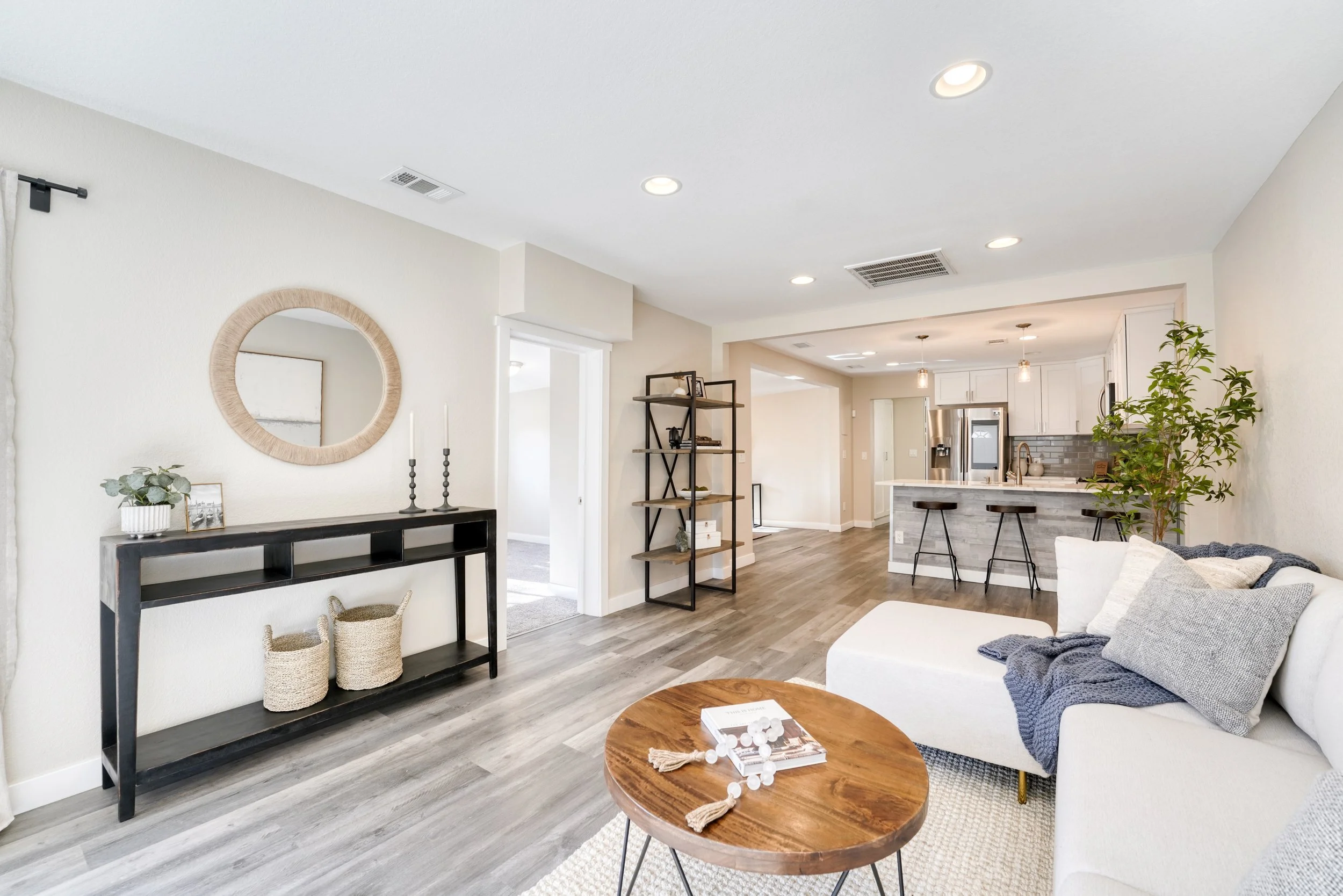 Open-concept living room and kitchen with white walls and ceiling, hardwood floors, a round wooden coffee table with books, a white couch with patterned pillows, a black console table with decorative items, a black metal shelving unit, and a large po