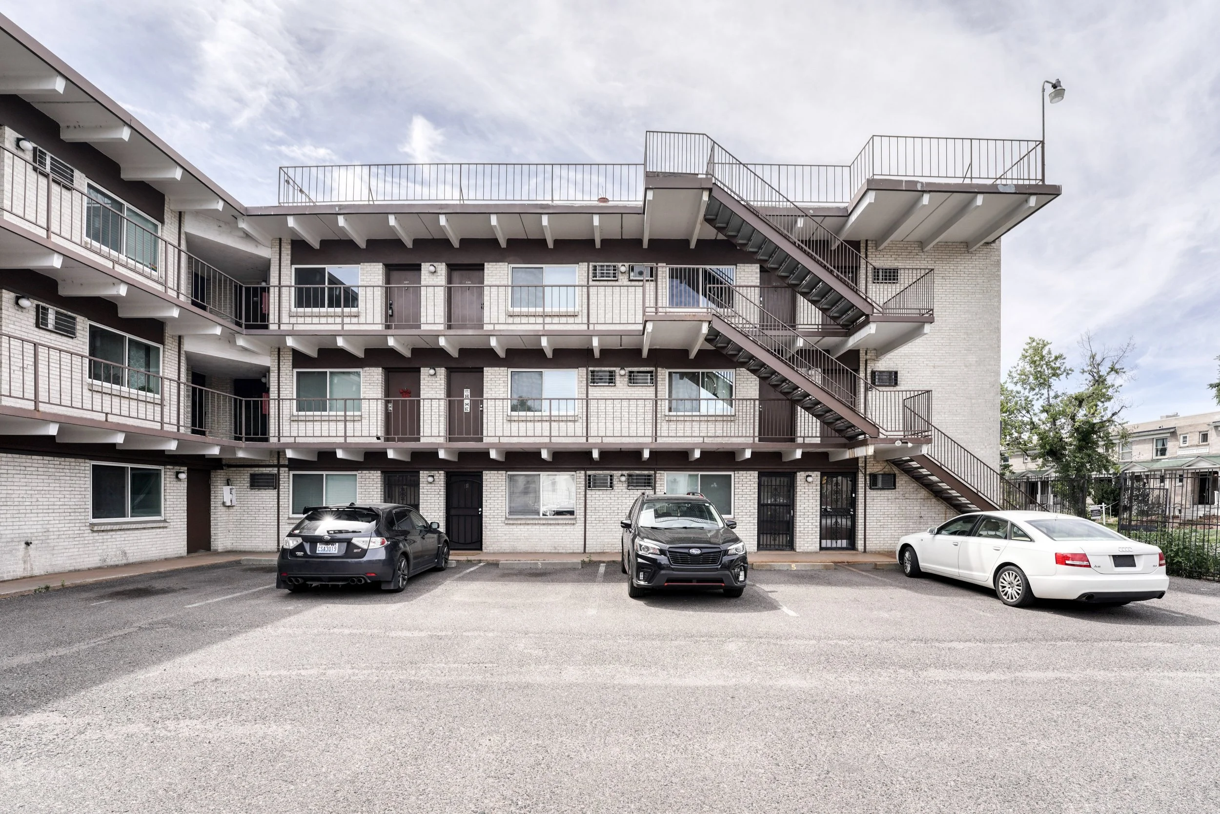 Four parked cars in front of a multi-story apartment building with external stairs and a parking lot under an cloudy sky.