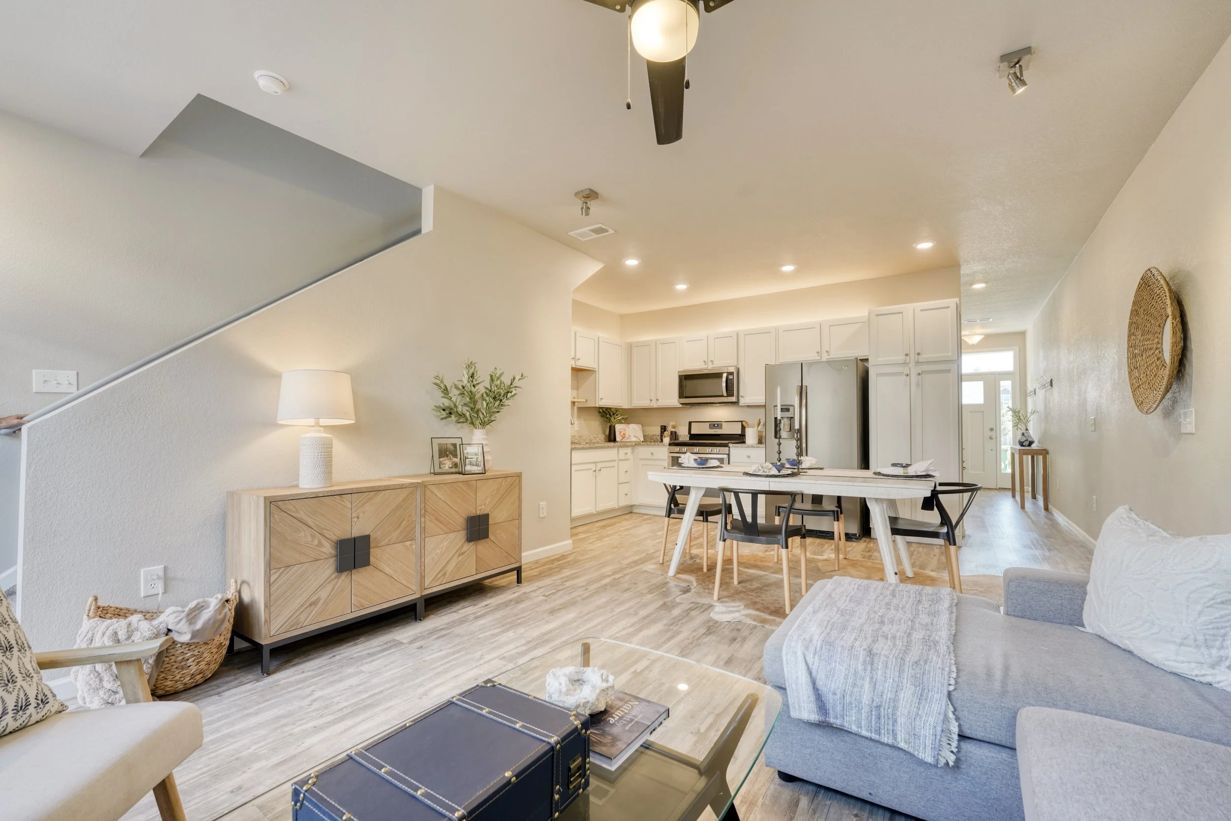 Open concept living room and kitchen with white cabinets, stainless steel appliances, and a wooden dining table with four chairs. The space features a neutral color palette with light walls and wood flooring.