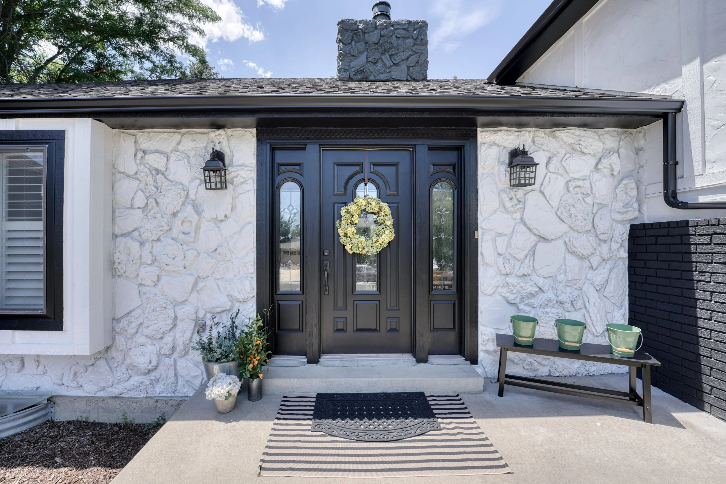 Front view of a house entrance with stone and white walls, black door with glass panels and floral wreath, two outdoor lantern lights, a black and white striped rug, potted plants, and green cups on a small table.