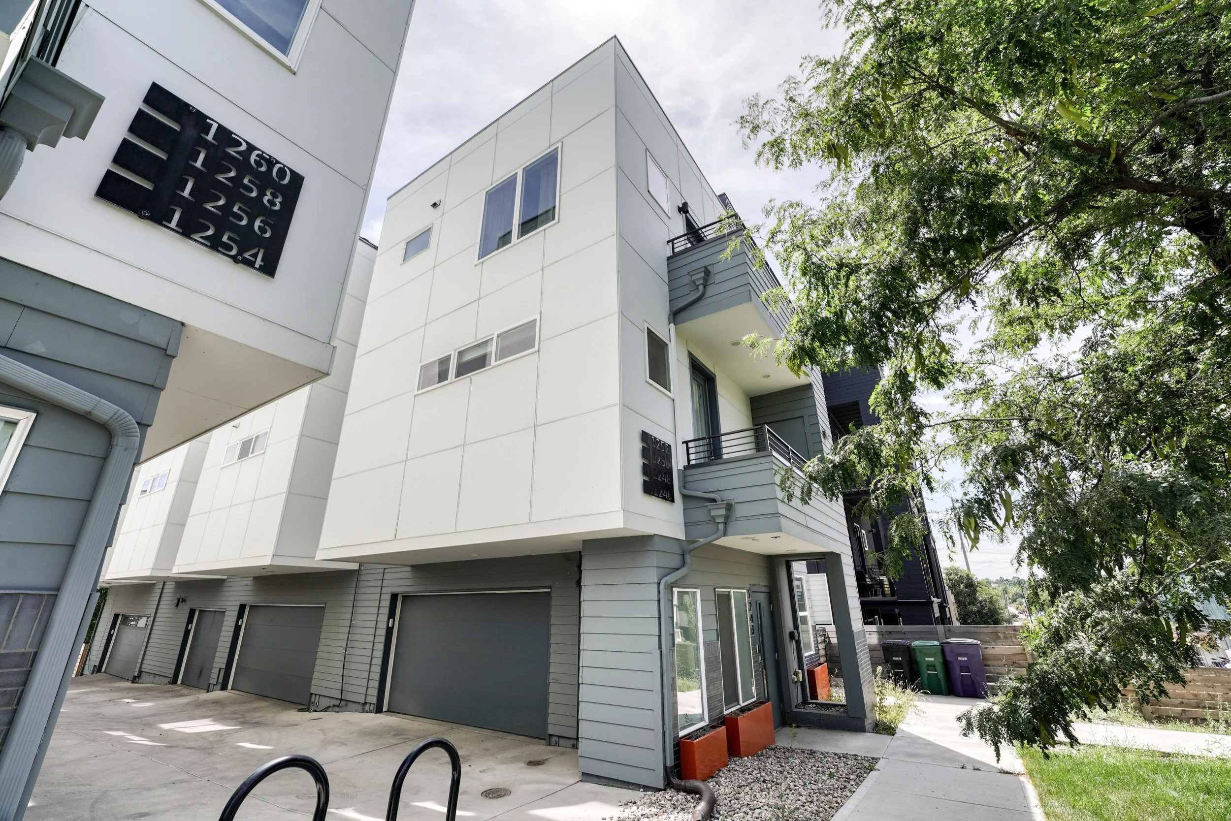 The image shows a modern multi-story residential building with a white exterior, several windows, and a small balcony. There is a tree with green foliage on the right side, and a driveway with garage doors and a sidewalk in front. The sky is partly c