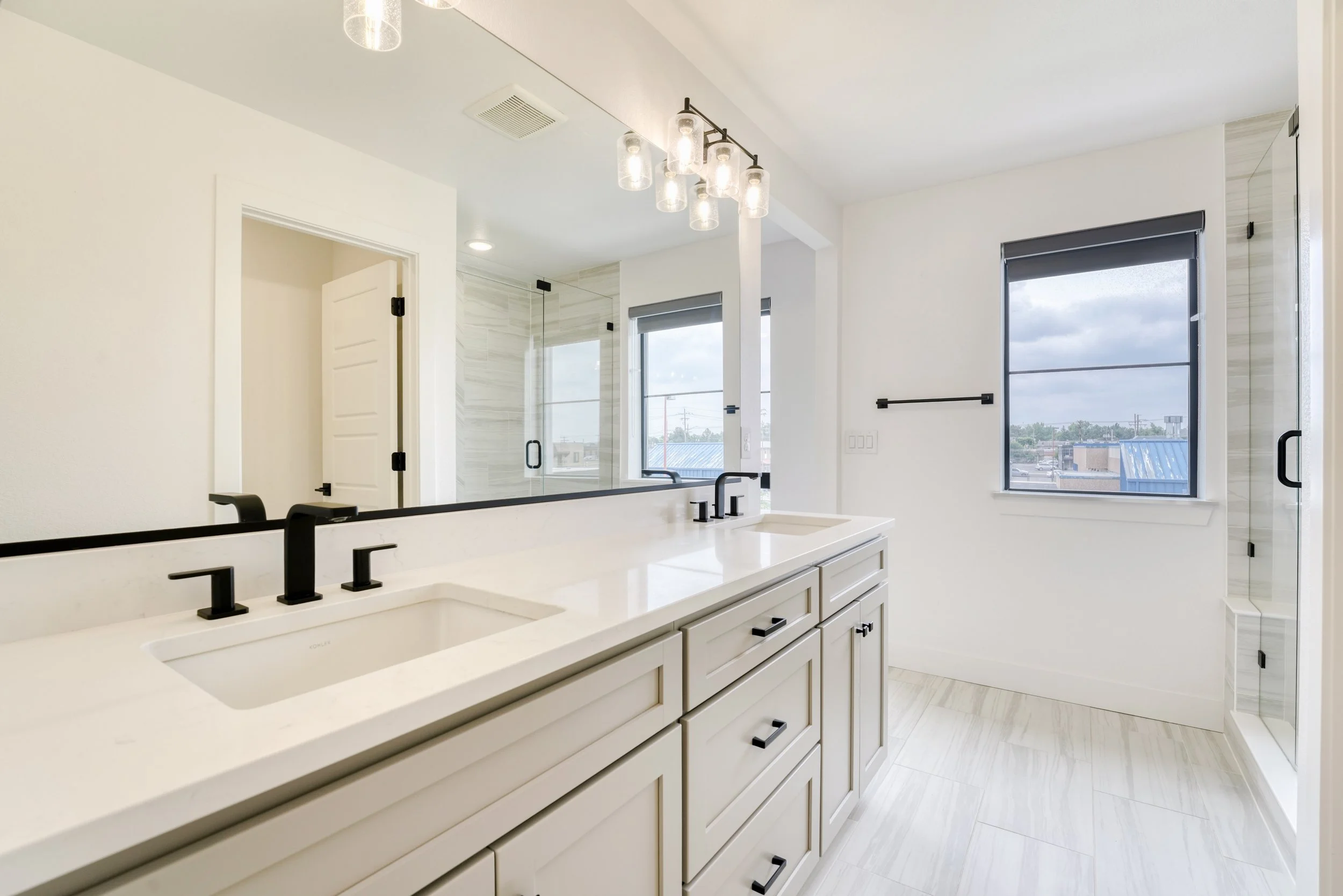 Modern bathroom with white cabinetry, black fixtures, and large windows showing a cloudy sky outside.