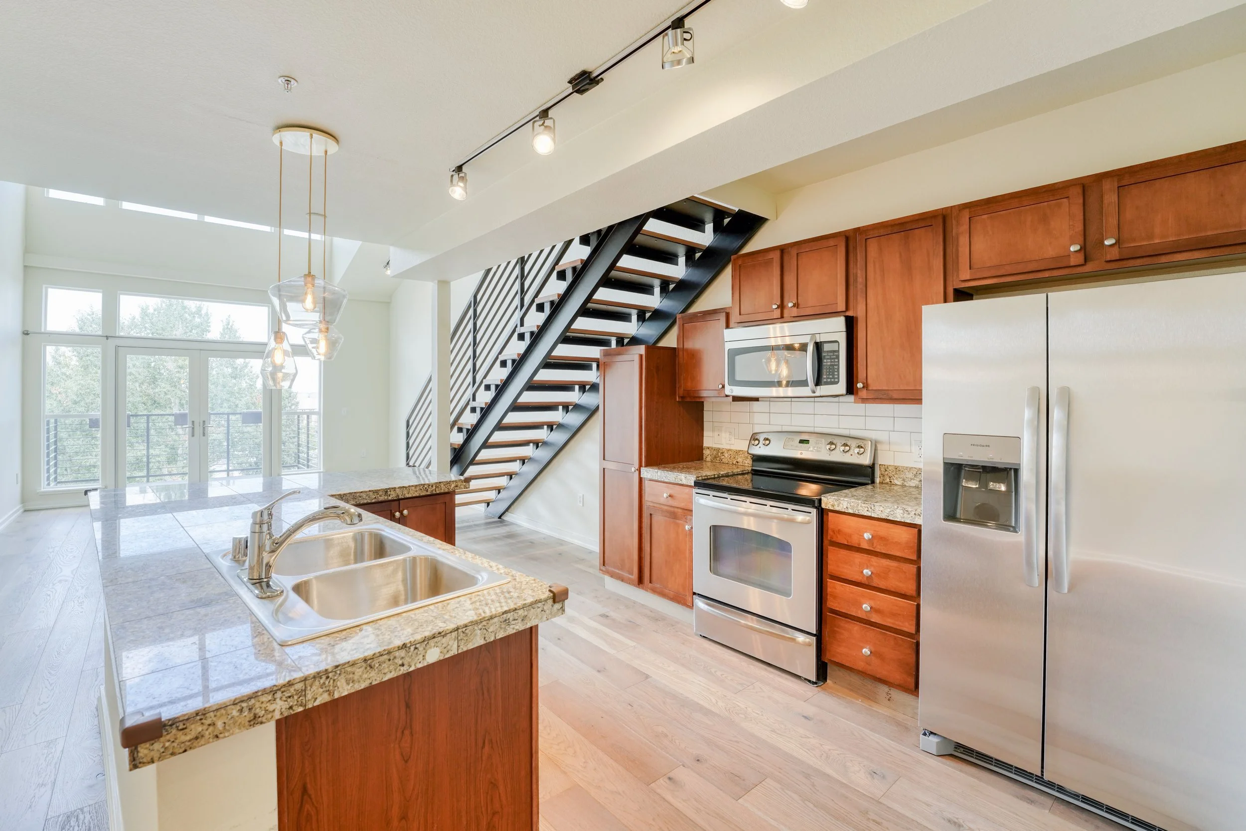 Modern kitchen with wooden cabinets, stainless steel appliances including a microwave and refrigerator, granite countertops, and a staircase with black metal railing leading upstairs. Large windows and glass door allow natural light into the space.