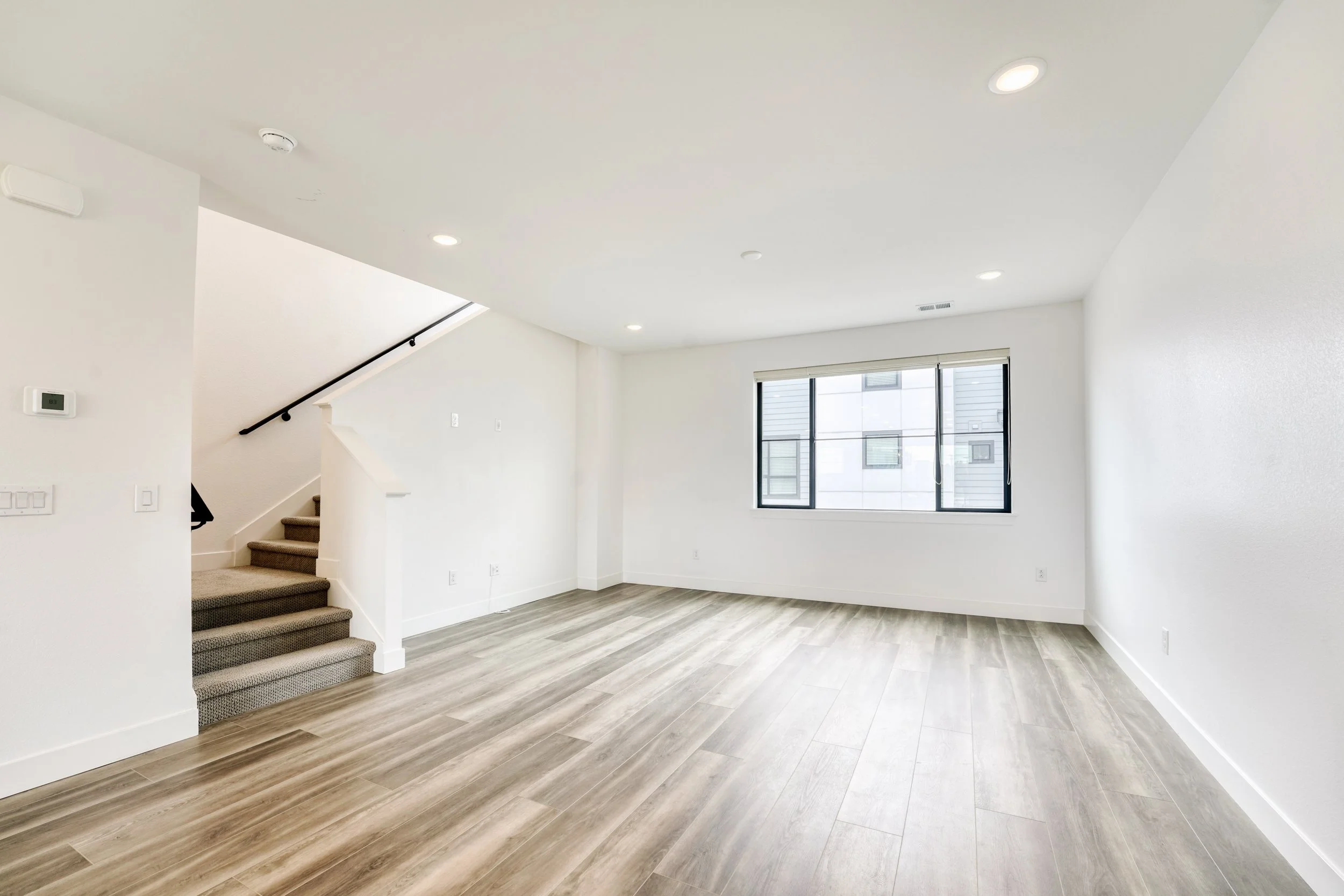 Empty living room with large window, white walls, wood flooring, and staircase with black handrail.
