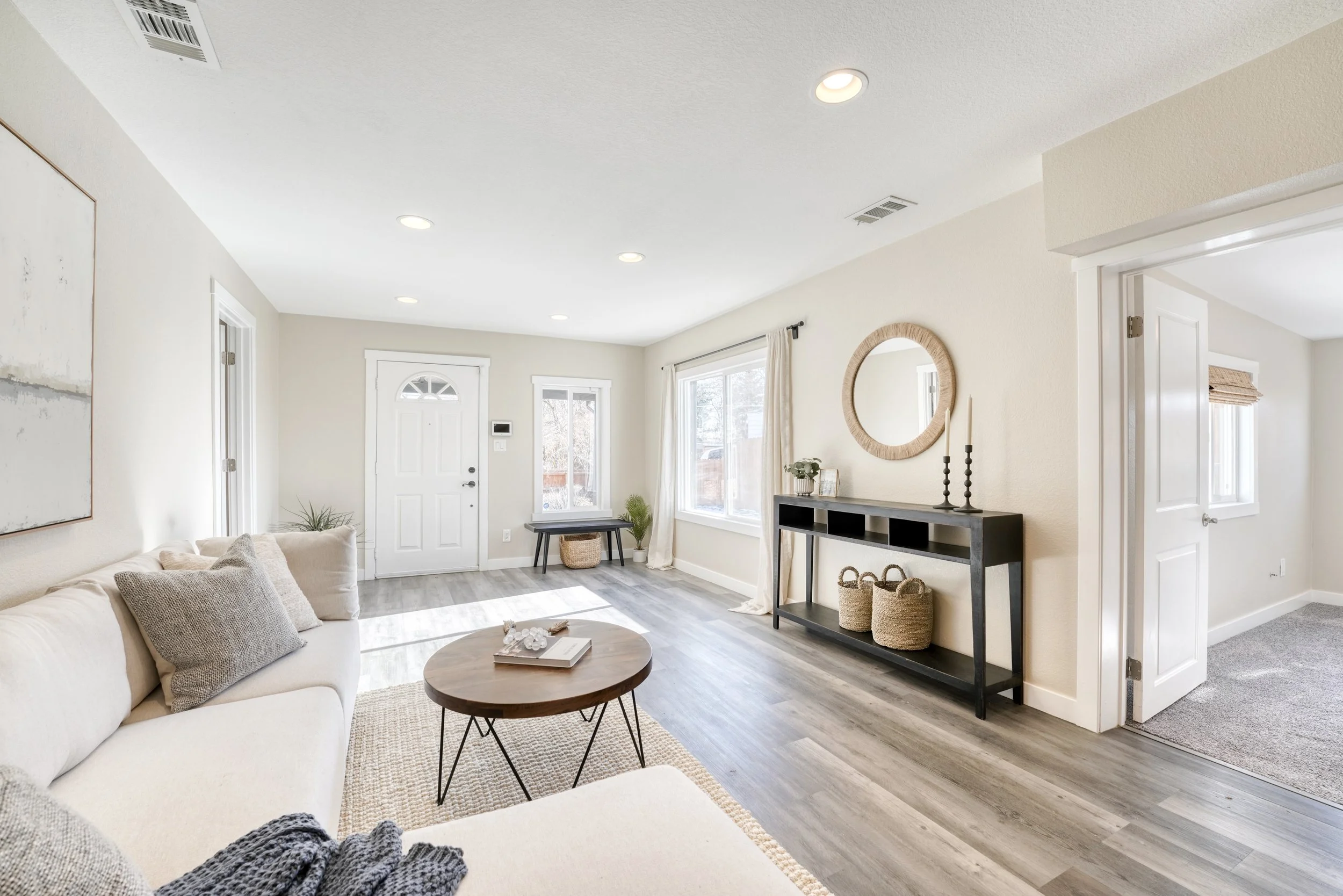 Living room with white sofa, round wooden coffee table, black console table with baskets, framed art on wall, and large windows with curtains.