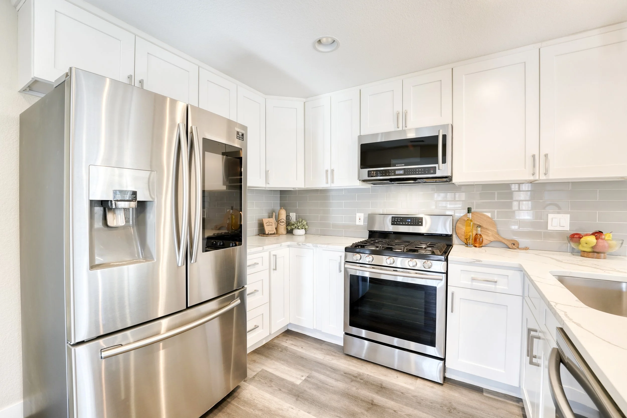 Modern kitchen with white cabinets, stainless steel refrigerator, microwave, and oven. Countertop with utensils, a bowl of fruit, and decorative items.