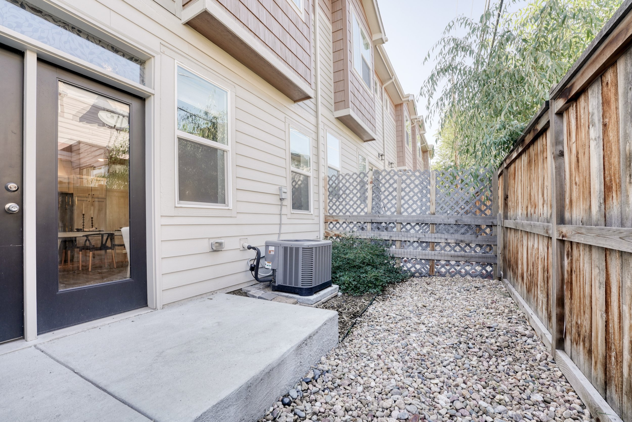 Backyard view of a house with a concrete patio, gravel ground, and a wooden privacy fence. Windows and a door to the house are visible, along with an air conditioning unit outside.