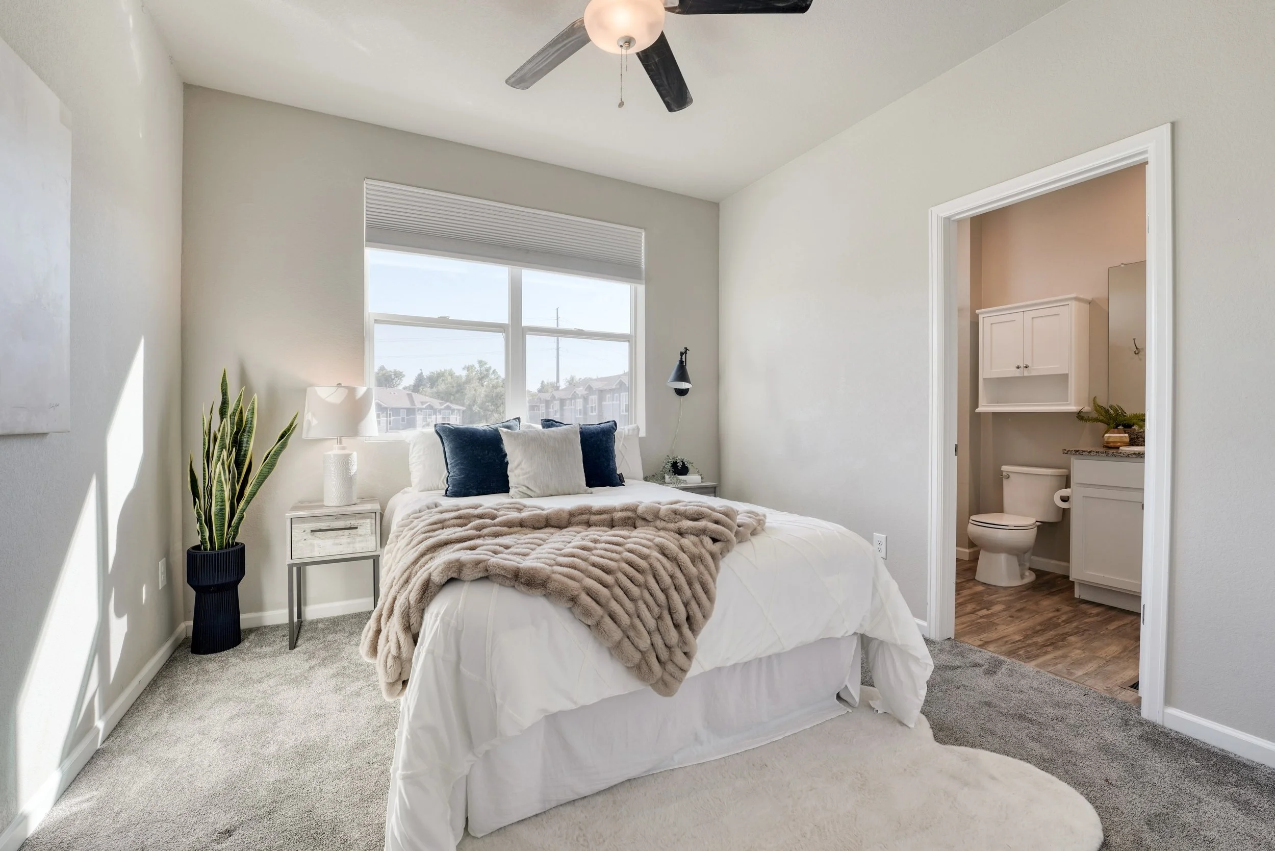 A bedroom with a white bed, beige blanket, and dark pillows. A side table with a white lamp, a potted plant, and a small decorative object. A window with a view of houses outside. A ceiling fan above. An adjoining bathroom with a toilet and white cab