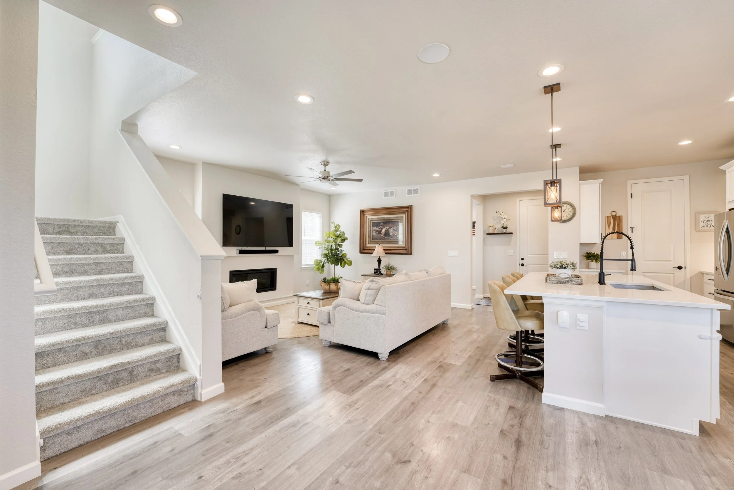 Open-concept living room and kitchen with light-colored furniture, a large flat-screen TV, a ceiling fan, potted plants, a framed painting, white cabinets, a kitchen island with beige barstools, pendant lighting, and hardwood flooring.
