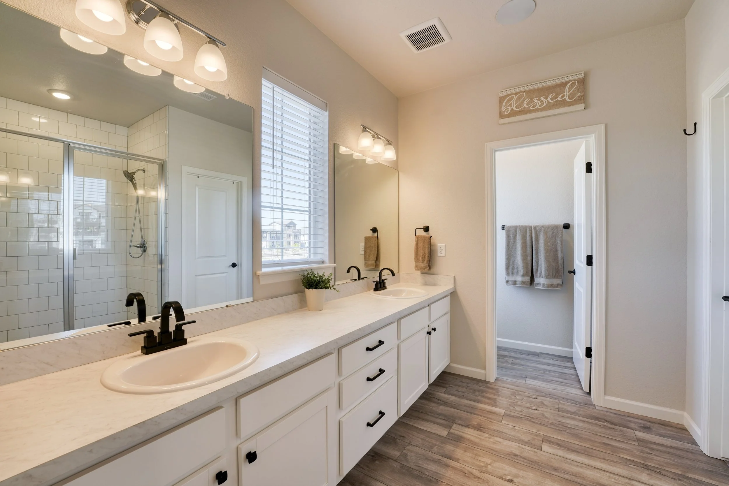 A modern bathroom with a large mirror above a white vanity with double sinks, black faucets, and drawers. The bathroom has a shower with white tile and a glass door, a window with white blinds, and a wooden sign that says 'blessed'. There are beige t