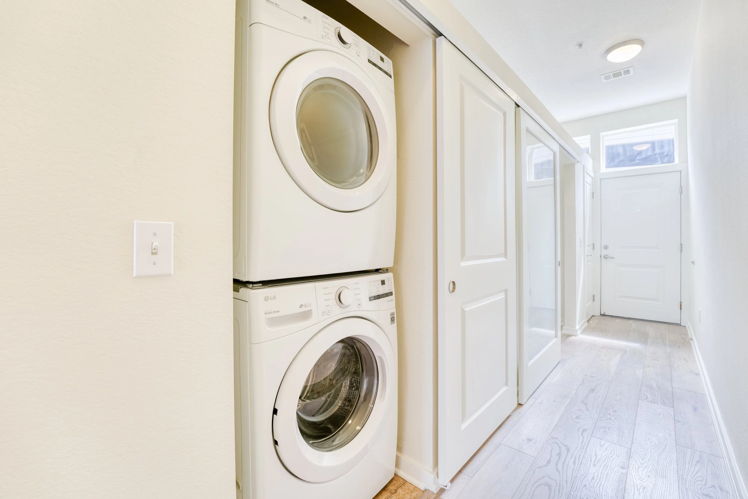 White stacked washer and dryer in a laundry closet with white cabinets and a hallway with a door, ceiling light, and wood flooring.