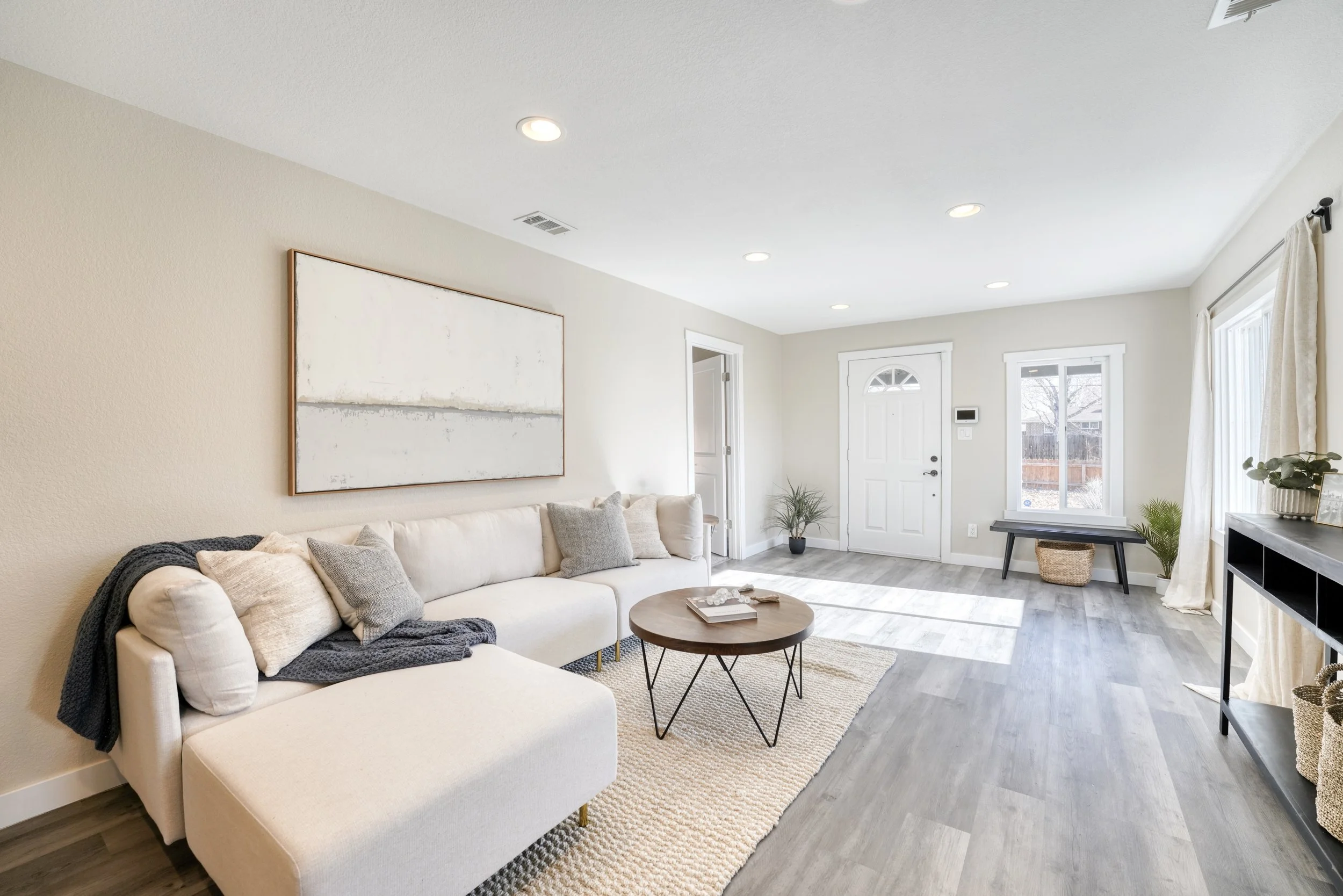 Bright living room with cream sofa, wooden coffee table, light-colored walls, windows with white curtains, and modern decor.