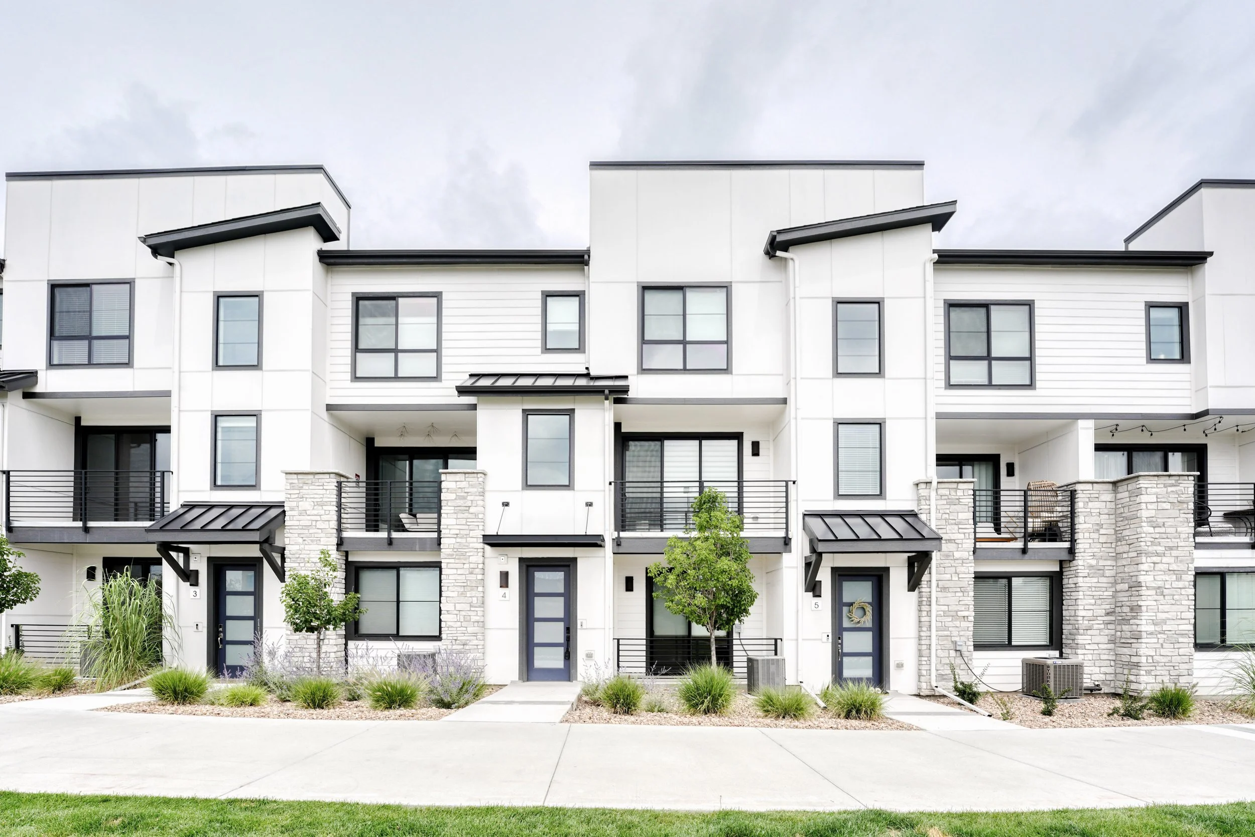 Front view of modern white apartment building with black accents, multiple balconies, and plants in the front yard.