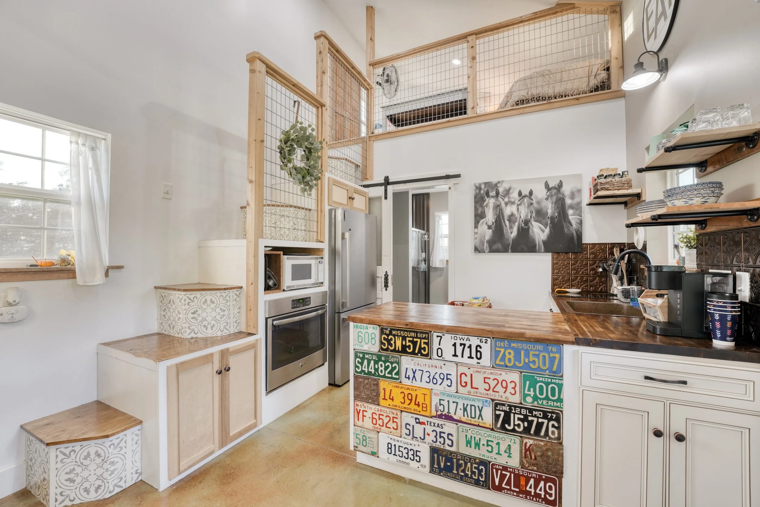 Interior view of a kitchen and living space with a loft. The kitchen features a refrigerator, microwave, and decorative license plates on a counter. The wall has a black and white horse photograph and open shelves with dishes. The loft area has a bed