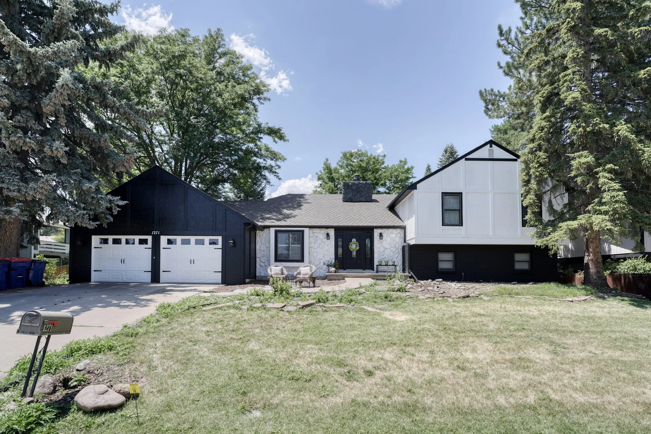 Front view of a modern two-story house with black and white exterior, a double garage, a small front porch with two armchairs, and large trees on a sunny day.