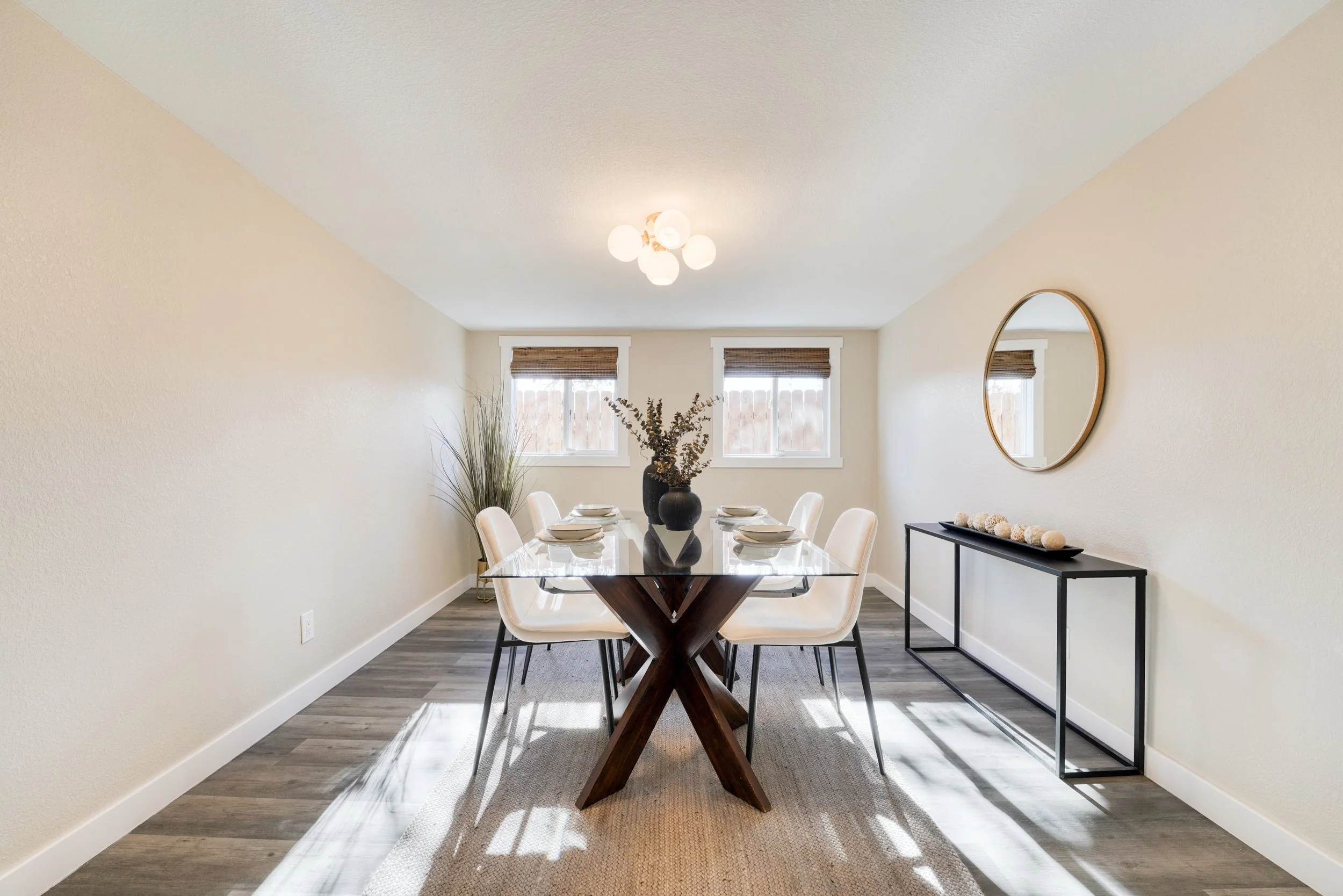 A dining room with a modern light fixture, a wooden table with a black base, six beige chairs, a black sideboard with decorative balls, a mirror on the wall, and two windows with bamboo blinds, natural light illuminating the space.