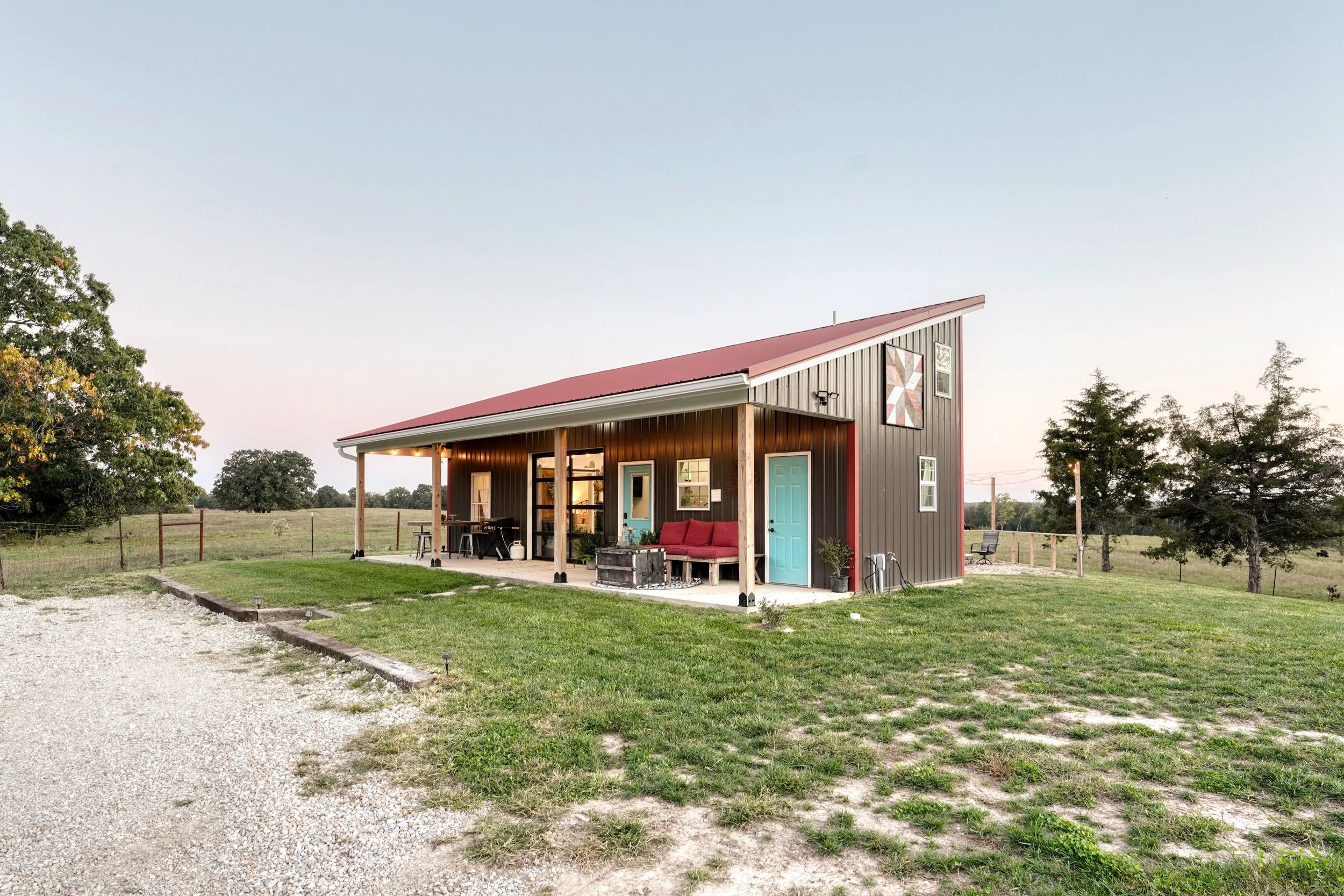 A modern barn-style house with black siding, a red sloped roof, and a light blue door, situated in a rural area with trees and open fields, during late afternoon or early evening.