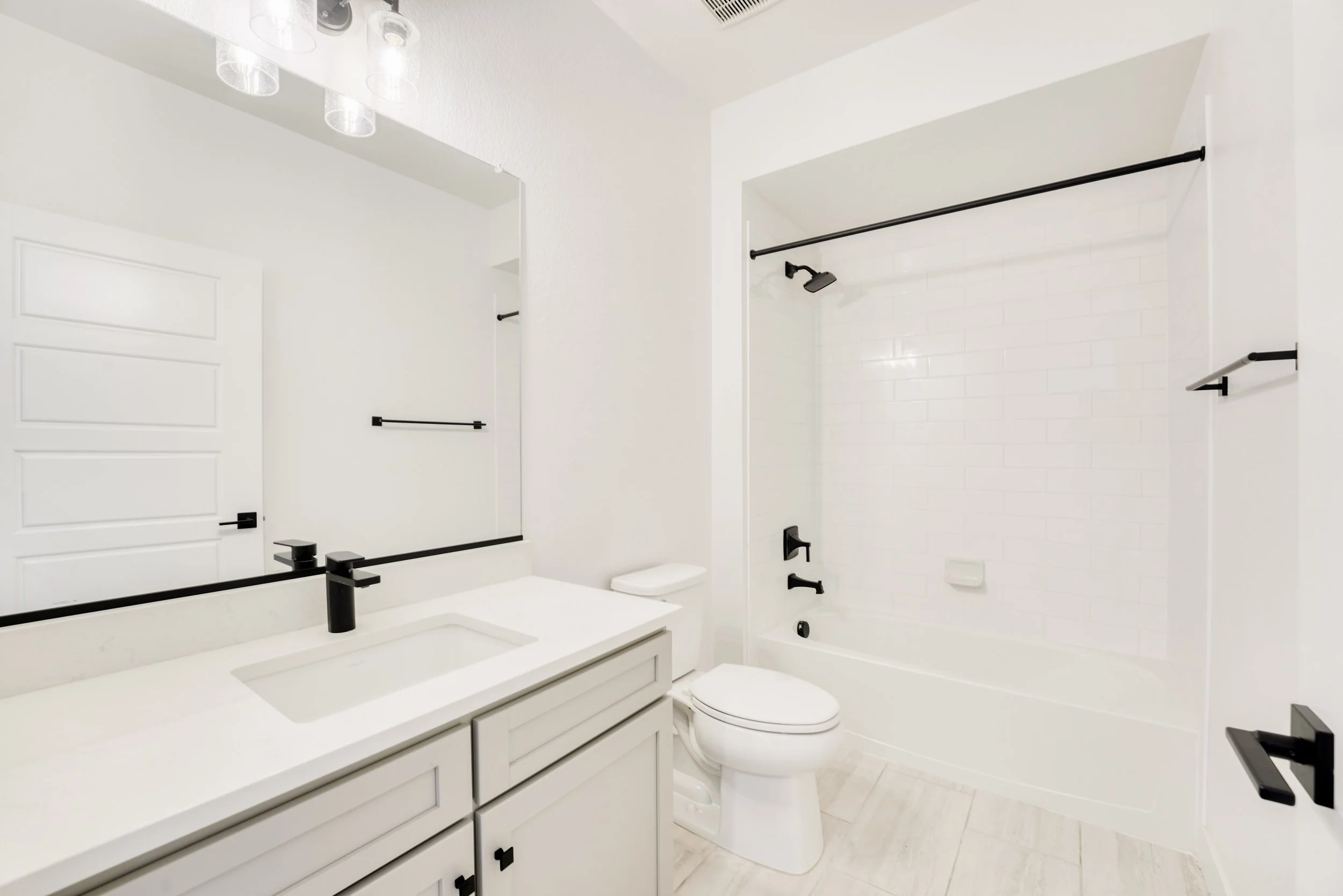 A modern, minimalist bathroom with white walls, a black faucet and fixtures, a large mirror, a white vanity with drawers, a white toilet, and a shower area with a black showerhead and a black curtain rod.