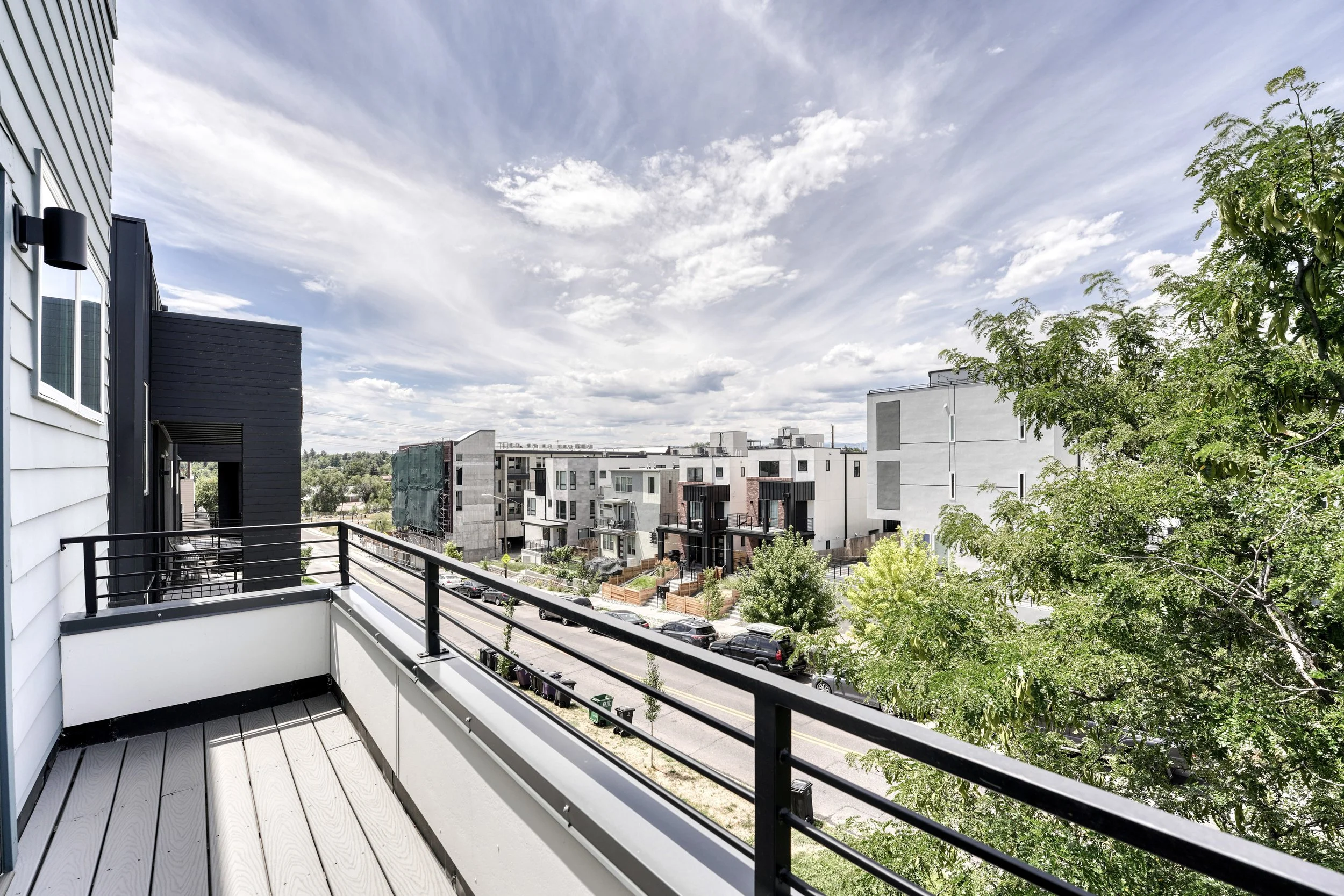 View from a balcony over a street with parked cars, modern buildings, and trees. The sky is partly cloudy with some blue visible.