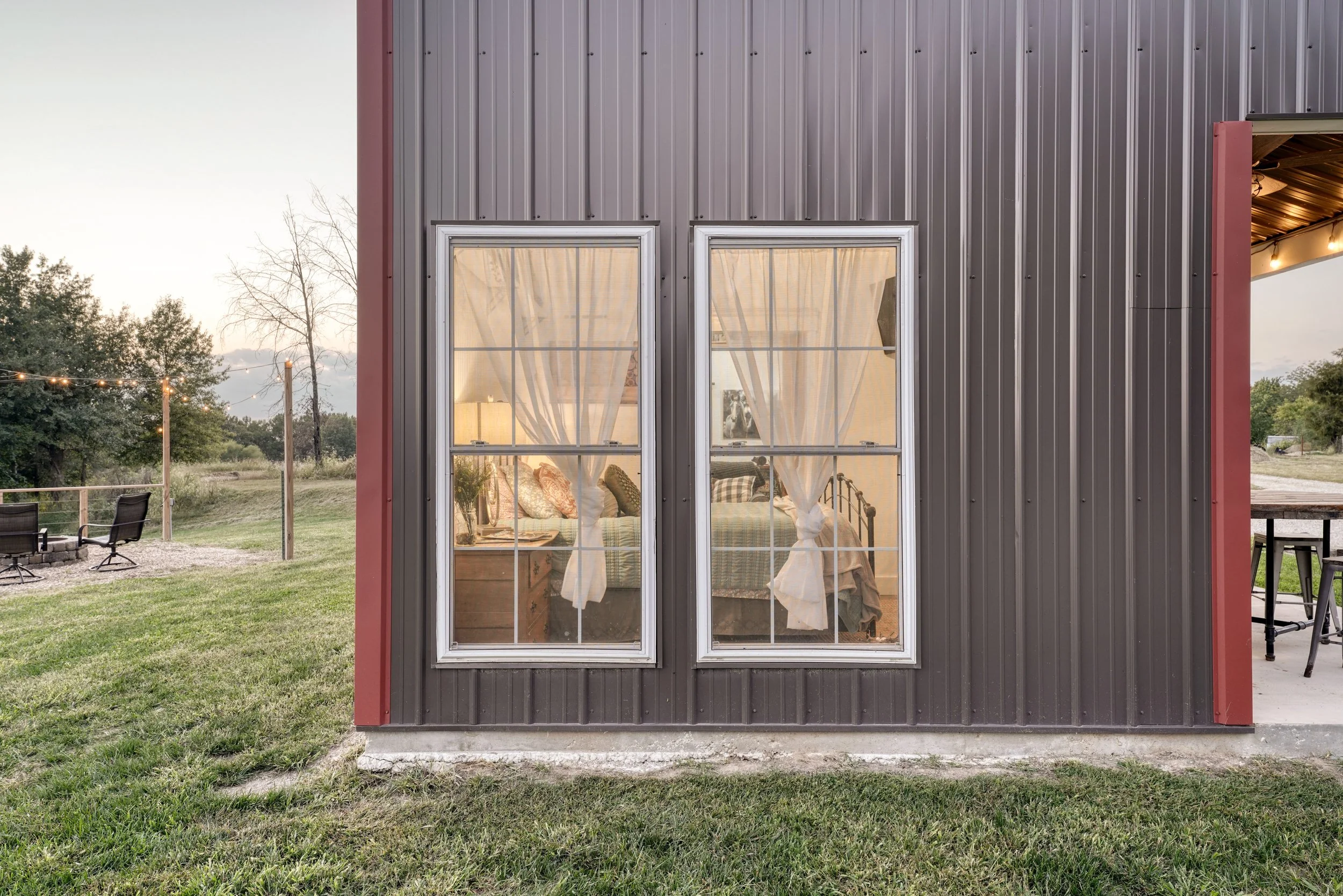 View of a building with metal siding, featuring two large windows with curtains, and a glimpse of a cozy interior bedroom with pillows. Outdoors, there's a grassy area with chairs and string lights, and trees in the background at sunset.