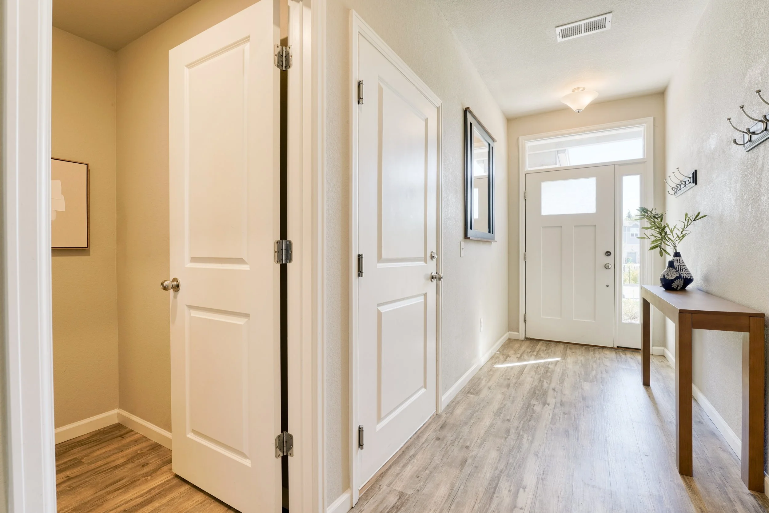 A bright entryway with a white front door and a small window above it. The floor is made of wood-like planks, and there is a small table with a decorative vase and plant. A wall-mounted coat rack with hooks is on the right wall, and there is a mirror