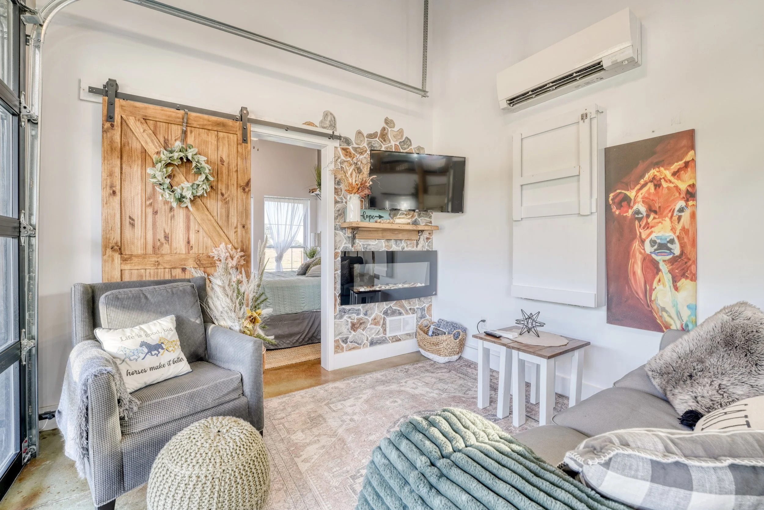 Cozy living room with a gray armchair, a knitted pouf, and a sofa with decorative pillows. There is a white side table, artwork of a bear, and a stone fireplace with a mounted TV. Natural light enters through a window, and the decor includes a wreath