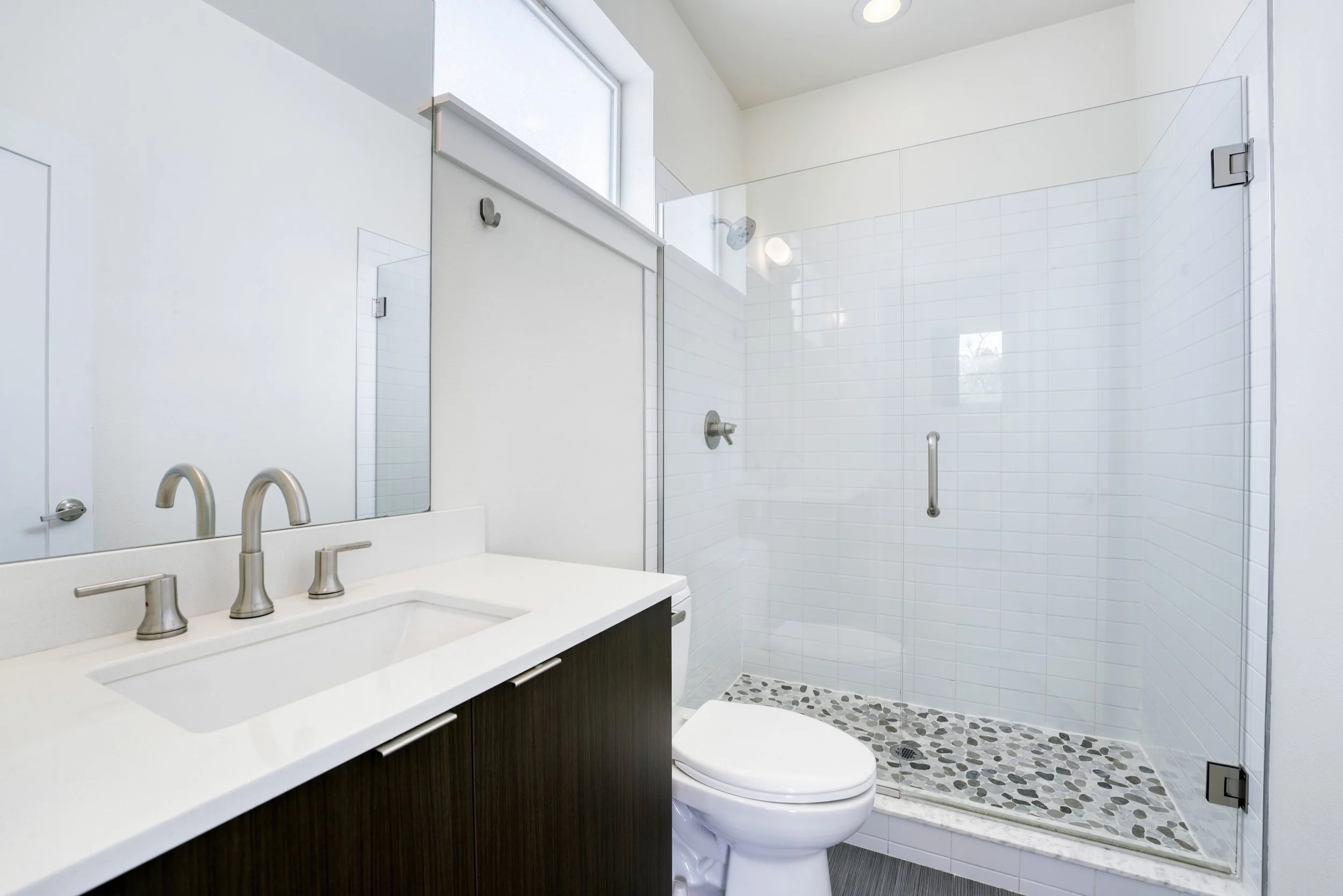 Modern bathroom with a white countertop sink and a dark wood vanity, a large mirror, a toilet, and a glass-enclosed shower with white tiles and pebble-patterned floor.