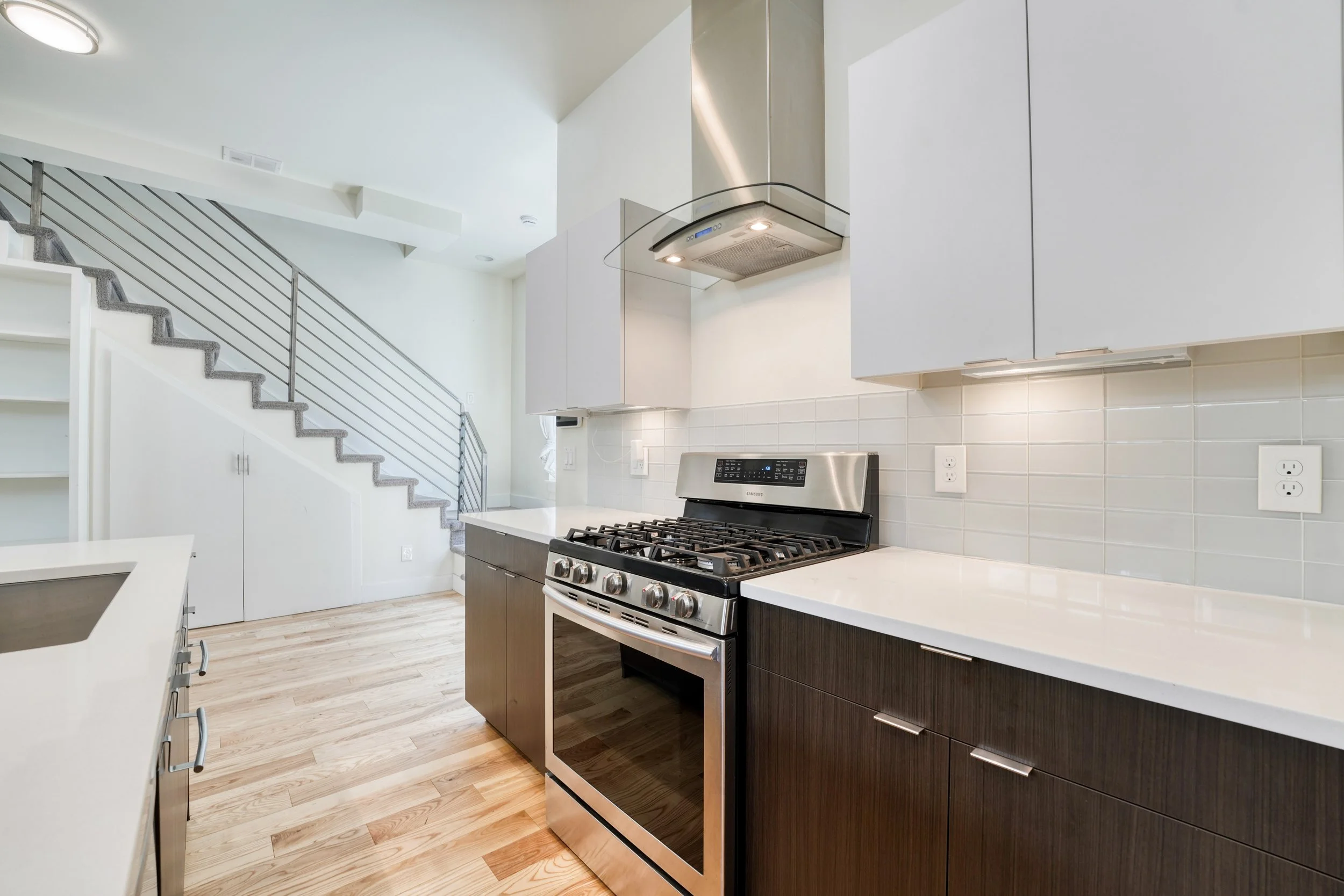 Modern kitchen with white countertops, dark wood cabinets, stainless steel stove, and white backsplash tiles. An over-the-range microwave and a staircase with metal railing are visible in the background.