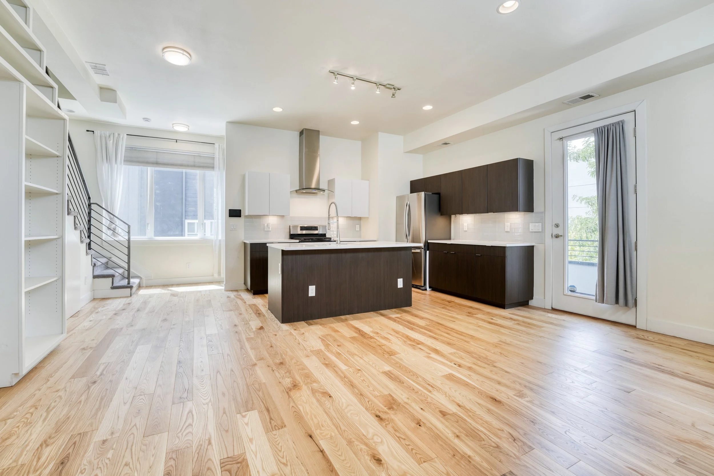 Modern kitchen with white and dark brown cabinets, stainless steel appliances, hardwood flooring, large window with white curtains, side door with a gray curtain, and vaulted ceiling with recessed lighting.