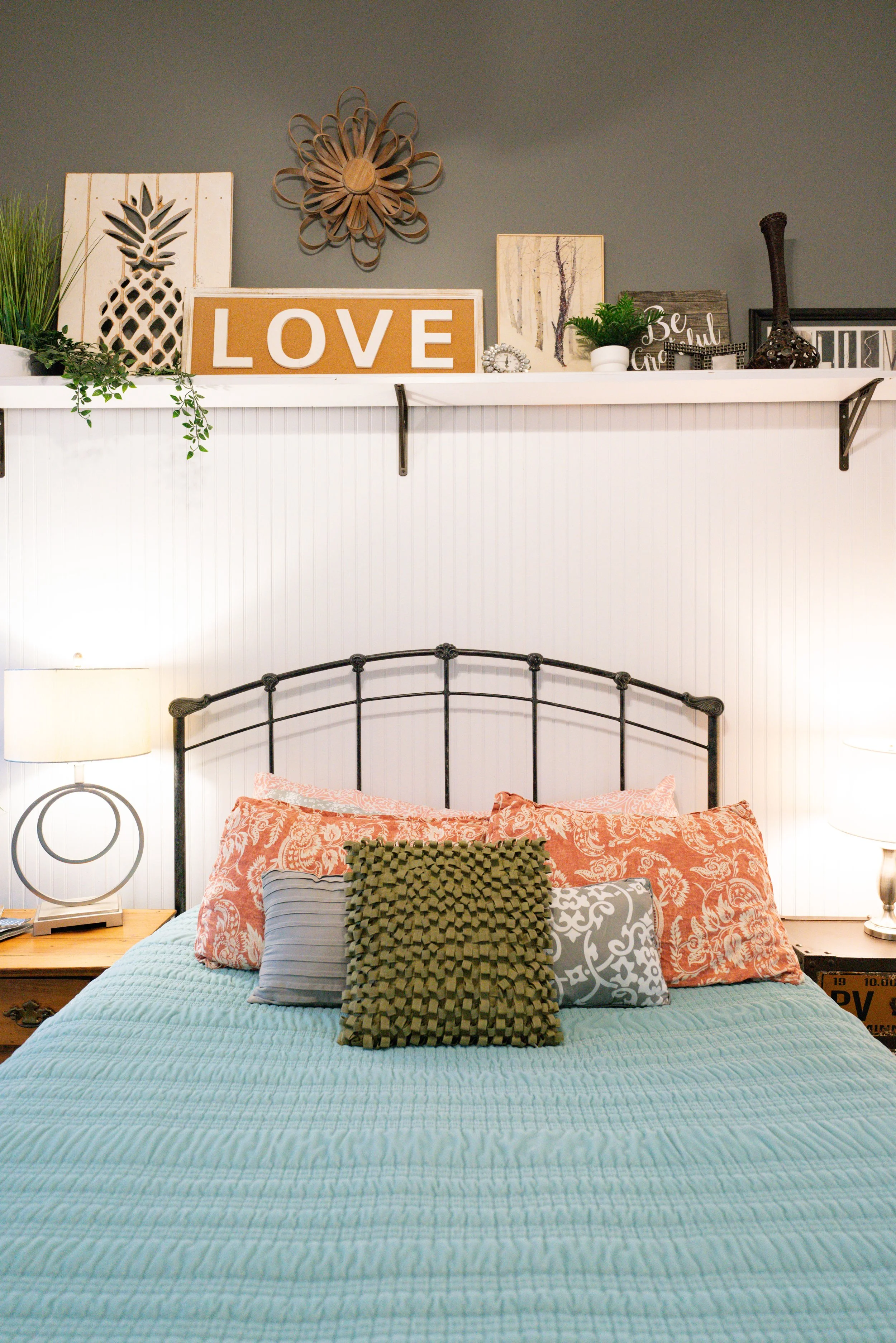 Bedroom with bed featuring a turquoise quilt and multiple pillows, nightstand with a lamp on either side, and a white shelf decorated with framed art, plants, and decorative objects above the bed.