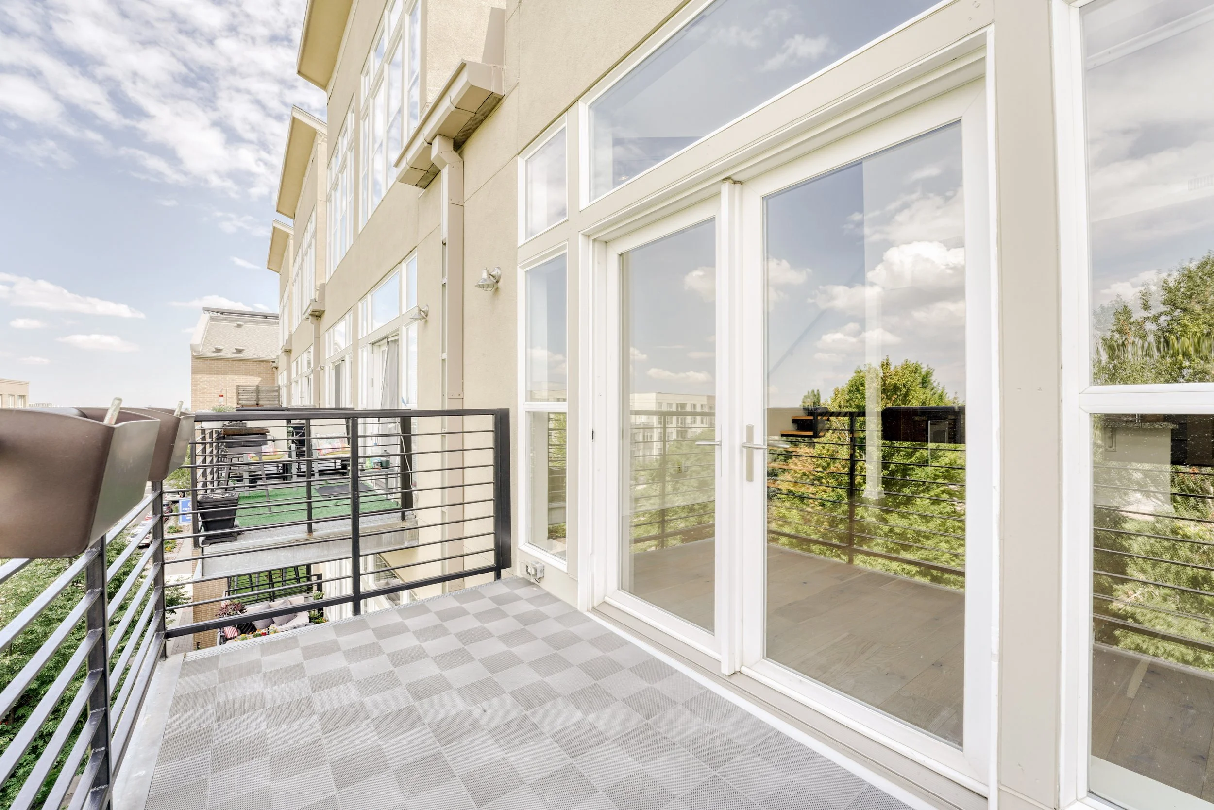 Balcony of a modern apartment building with glass sliding doors, a checkered floor, metal railing, and some outdoor furniture. Cityscape and trees can be seen in the background.