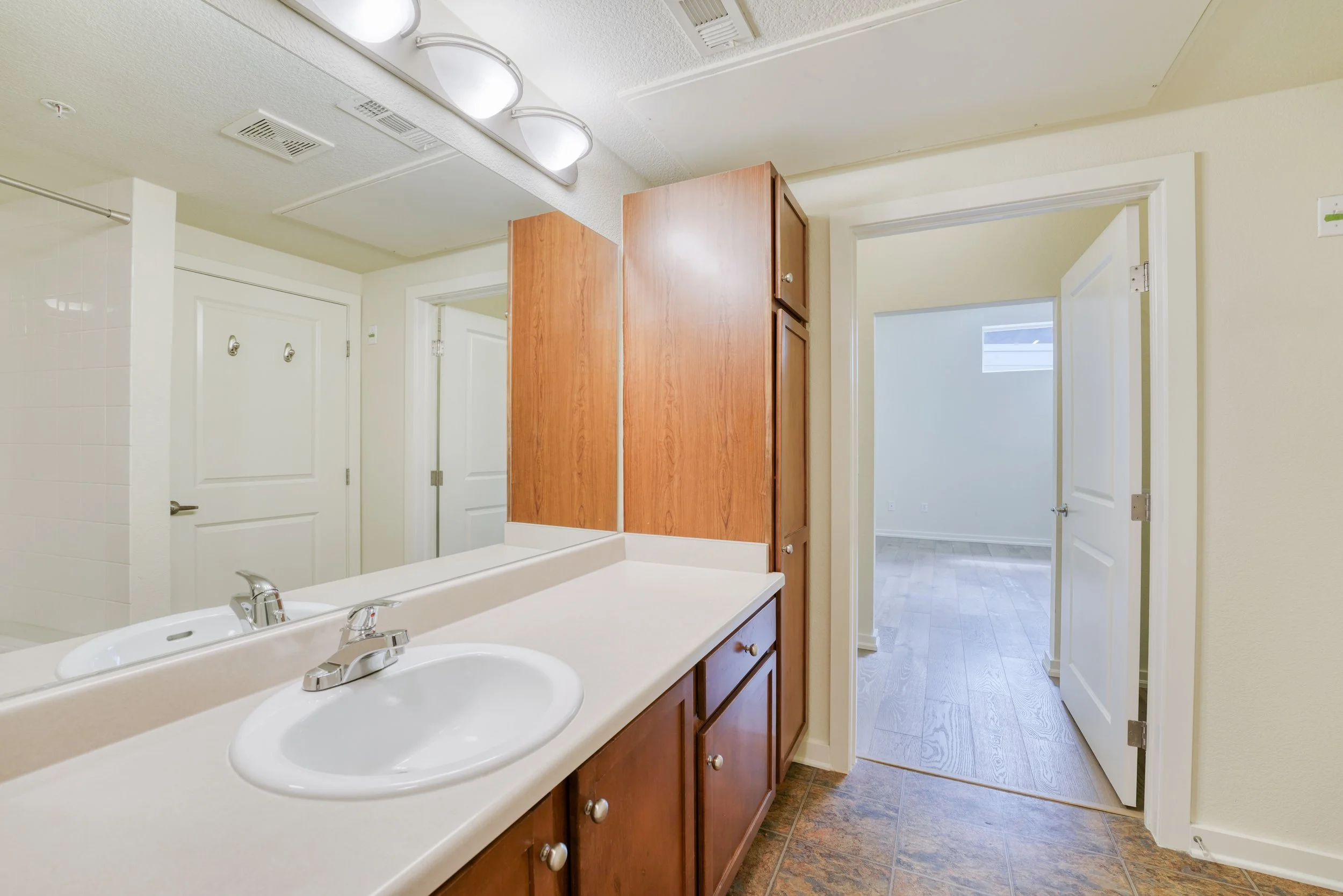 Bathroom with a double sink vanity, wooden cabinets, and a mirror. There is a doorway leading to another room with light-colored flooring and a small window.