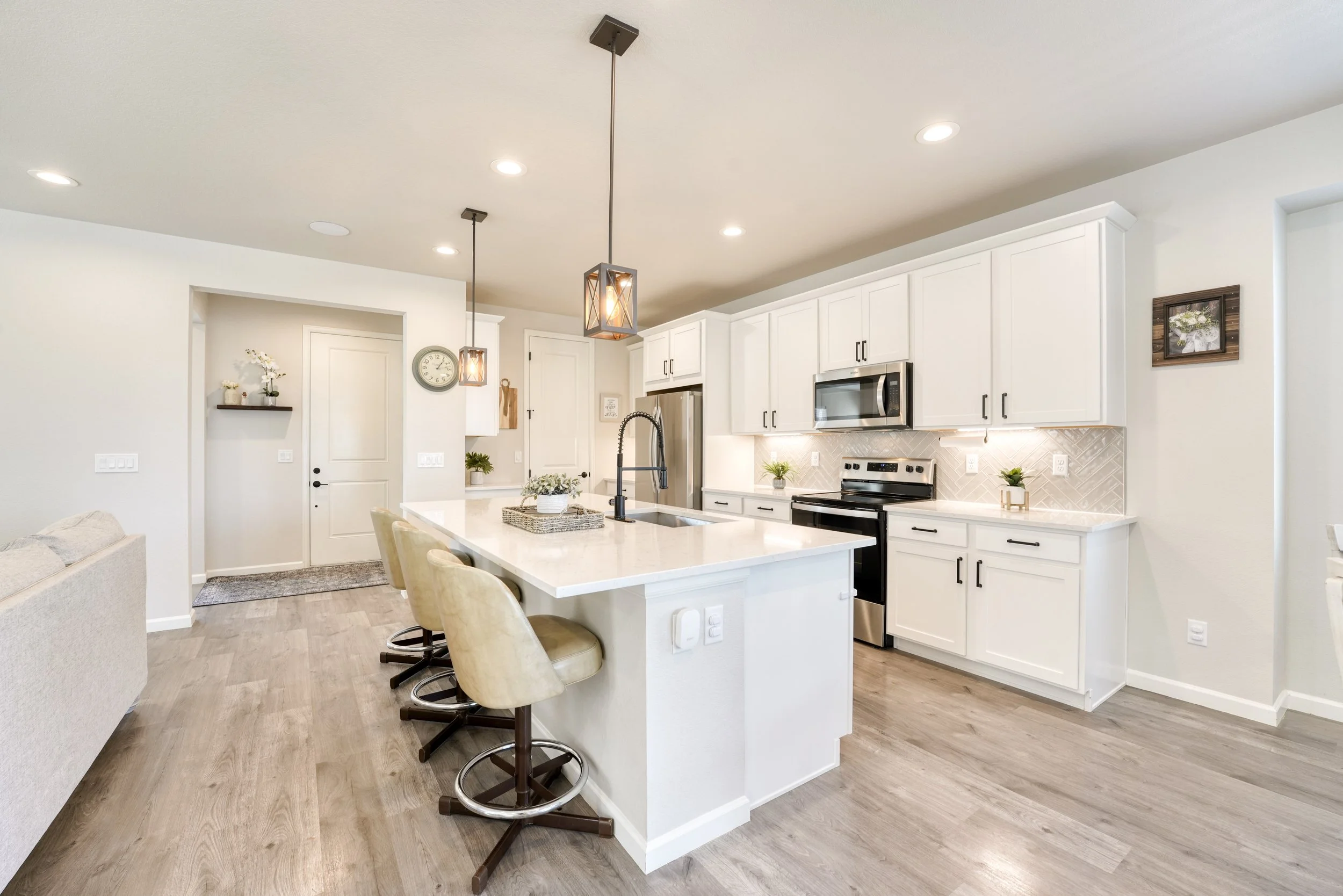 Modern kitchen with white cabinets, a large island with beige barstools, stainless steel appliances, pendant lights, and decorative plants.