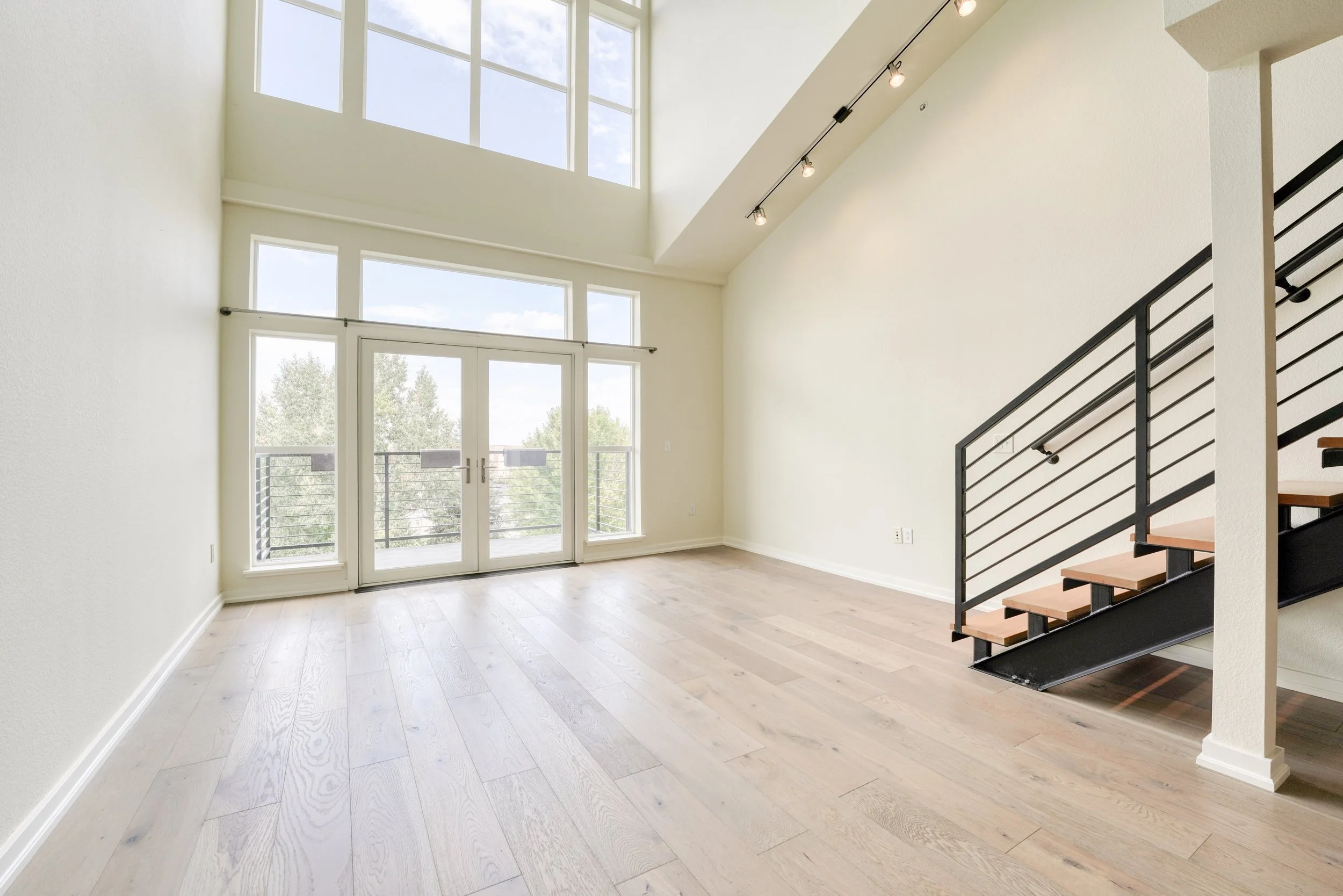 Bright interior of a modern living space with large windows, a balcony, light-colored hardwood floors, and a staircase with black metal railings.