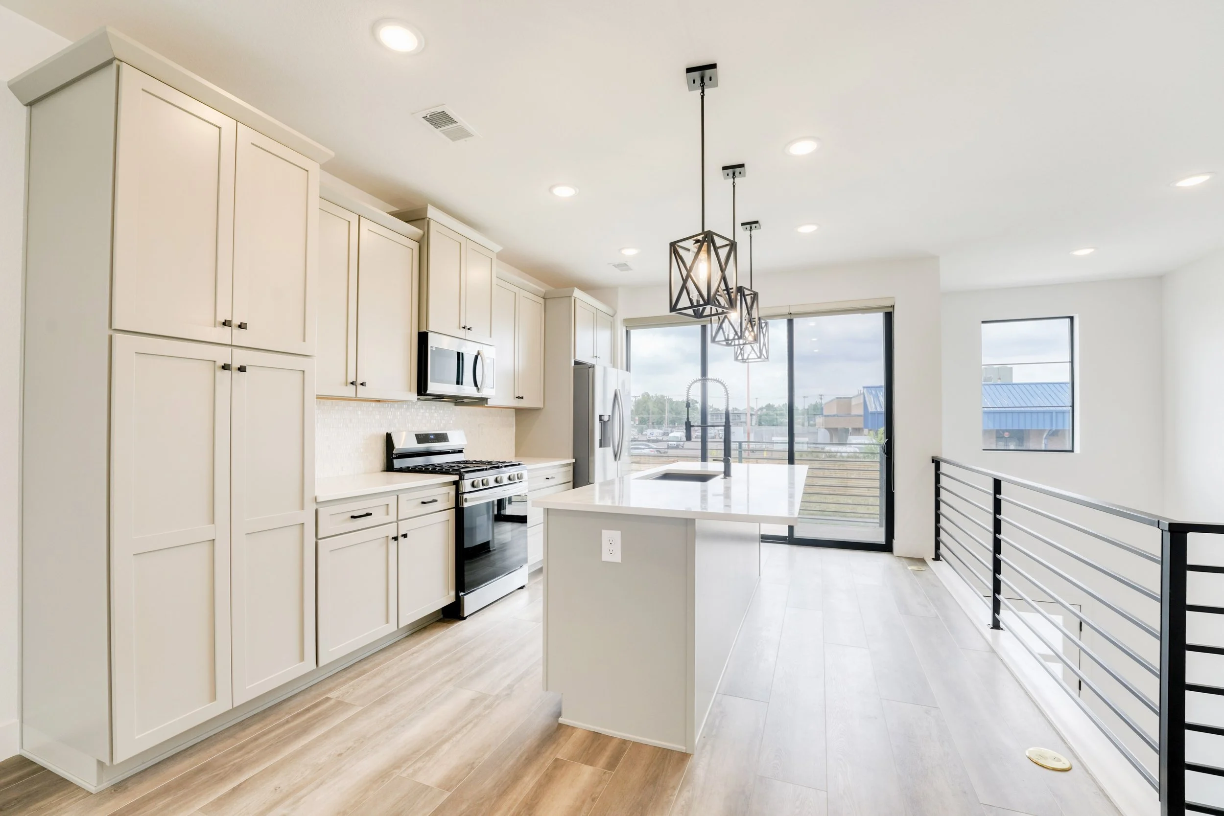 Modern kitchen with cream-colored cabinets, stainless steel appliances, a large window overlooking rooftops, pendant lighting, and light wood flooring.