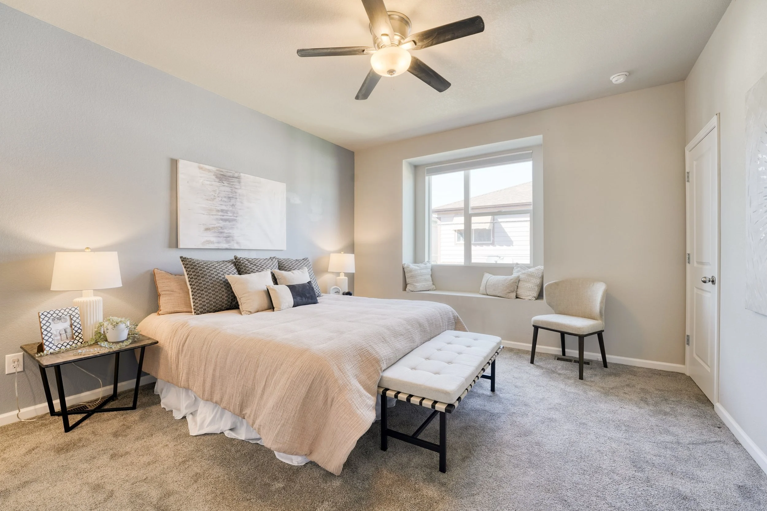 A bedroom with a bed, bedside tables with lamps, a window seat with cushions, and a ceiling fan. The room has light-colored walls and beige bedding.
