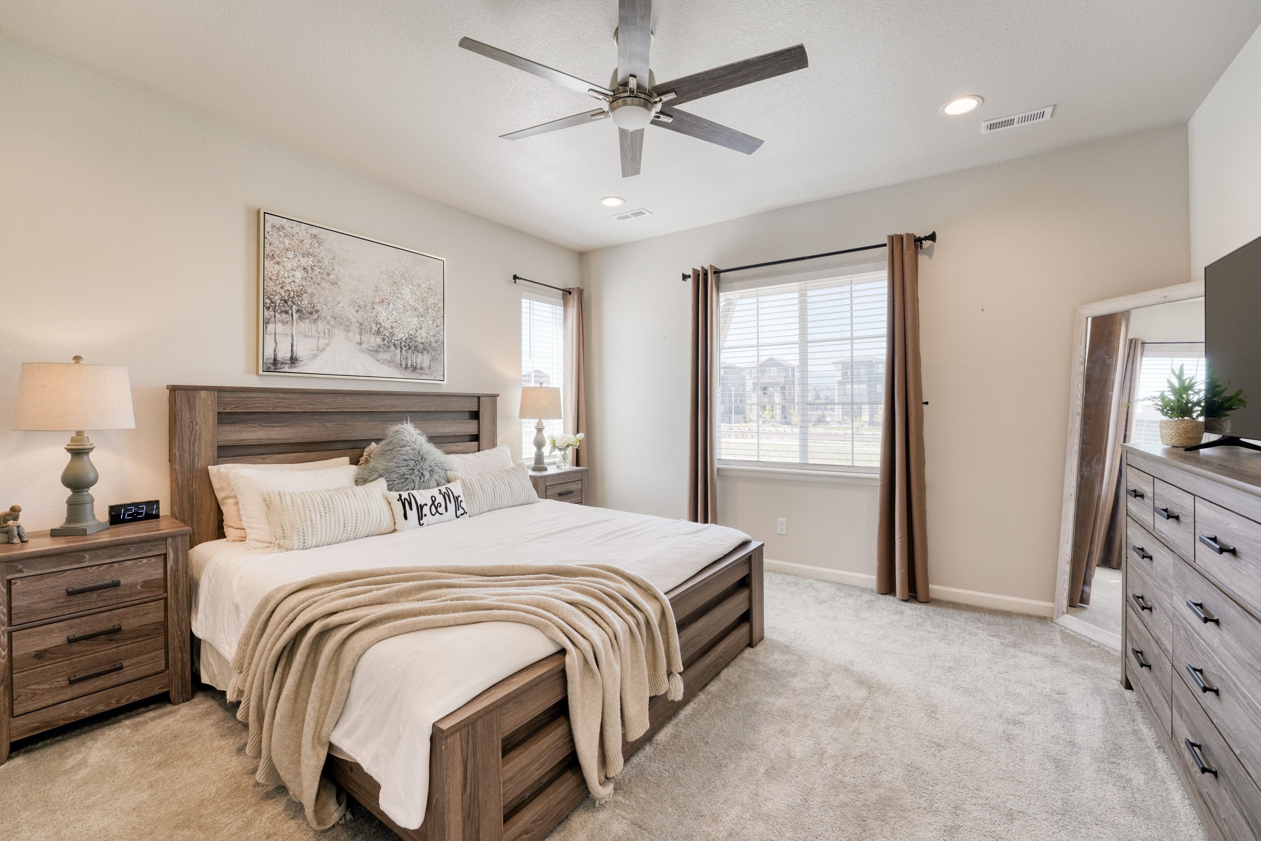 A cozy bedroom with a wooden bed frame, bedding, and decorative pillows. There are two nightstands with lamps, a dresser, and a mirror. The room has two windows with curtains and a ceiling fan.