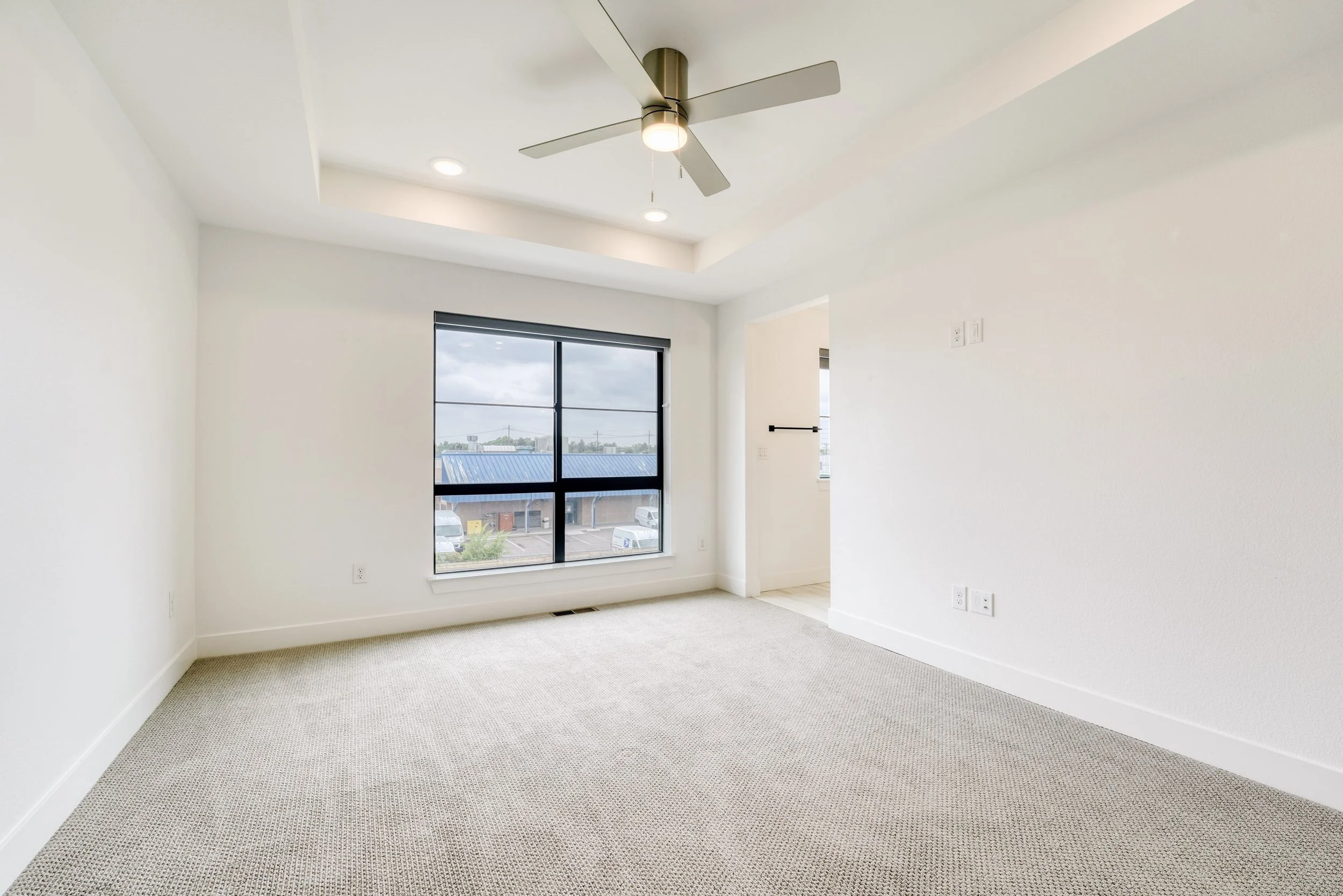 Empty room with white walls, large window, ceiling fan, and beige carpet.
