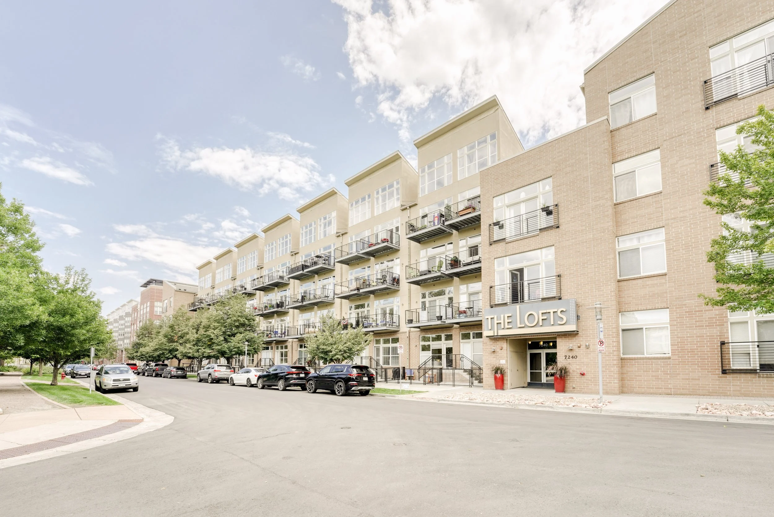 A multi-story residential building named The Lofts with balconies and rental signage, surrounded by a parking lot with several cars and trees, under a partly cloudy sky.