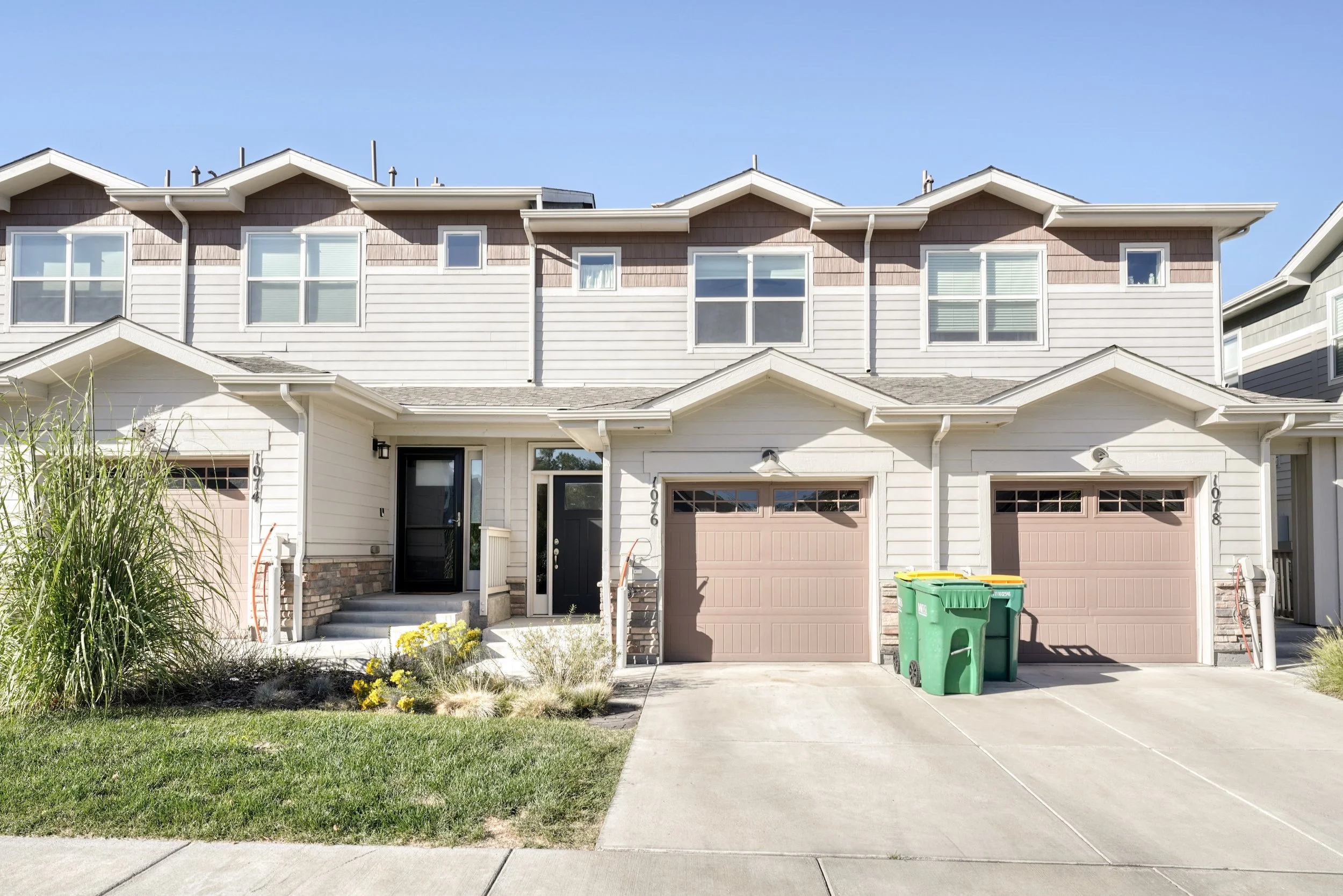 A row of modern townhouses with garages, a small front yard, and trash bins in the driveway, under a clear blue sky.