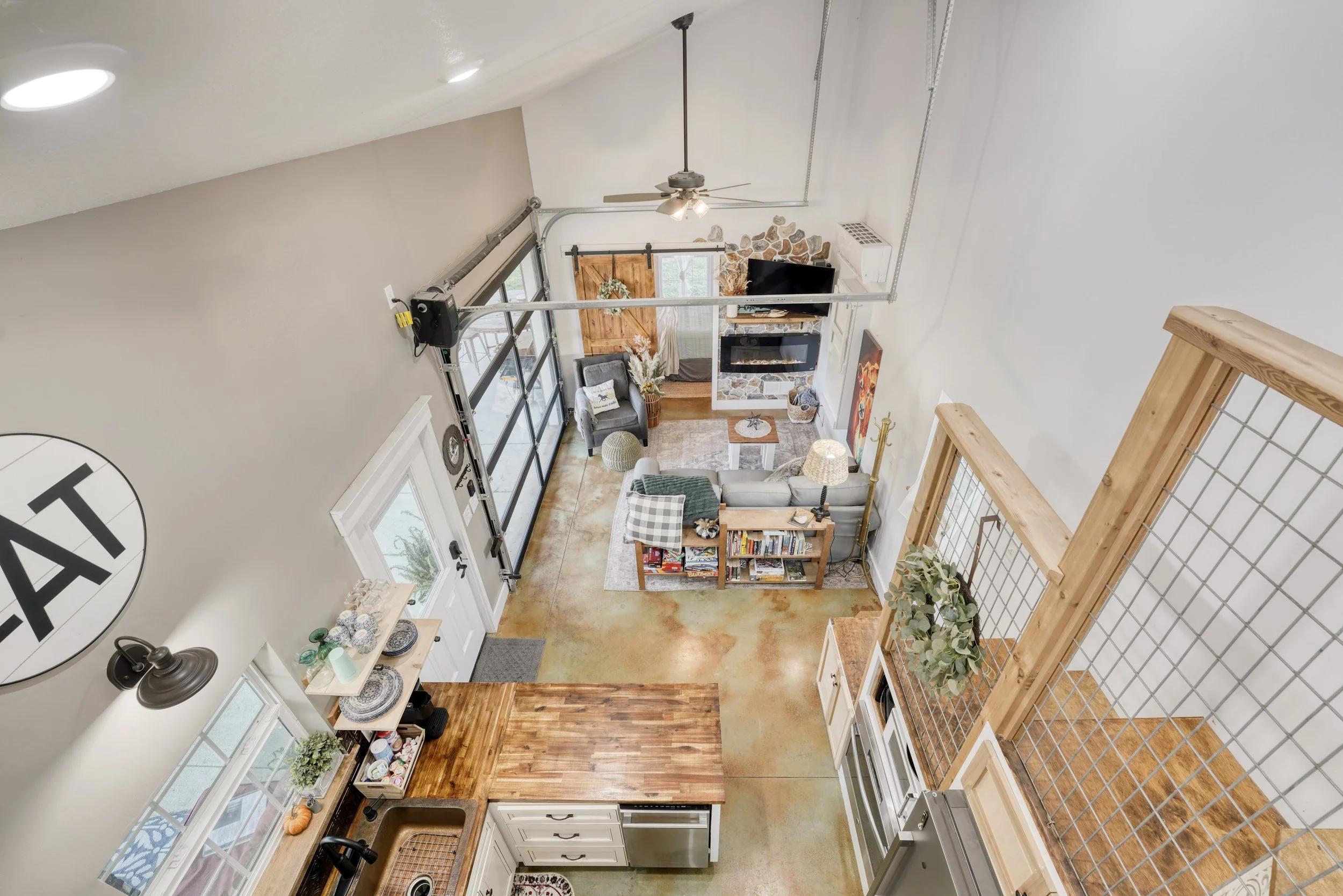 A top-down view of a spacious living room and kitchen area with a vaulted ceiling, ceiling fan, stone fireplace, flat-screen TV, sliding glass doors, and various furniture and decorative items.