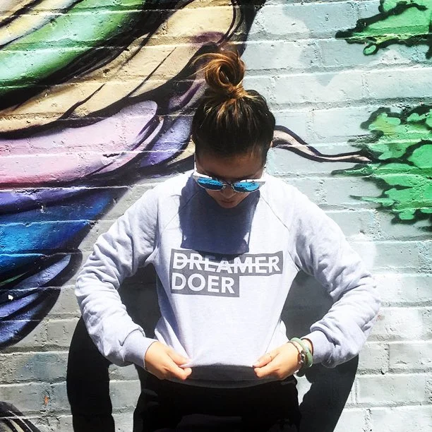 Person with a bun hairstyle and sunglasses standing in front of a colorful graffiti wall, wearing a white sweatshirt with the words 'DREAMER DOER' printed on it.