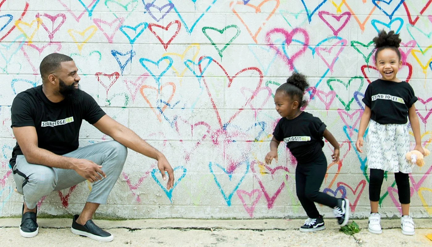 A man squatting and smiling at two young girls who are standing and jumping in front of a colorful wall painted with multiple hearts.