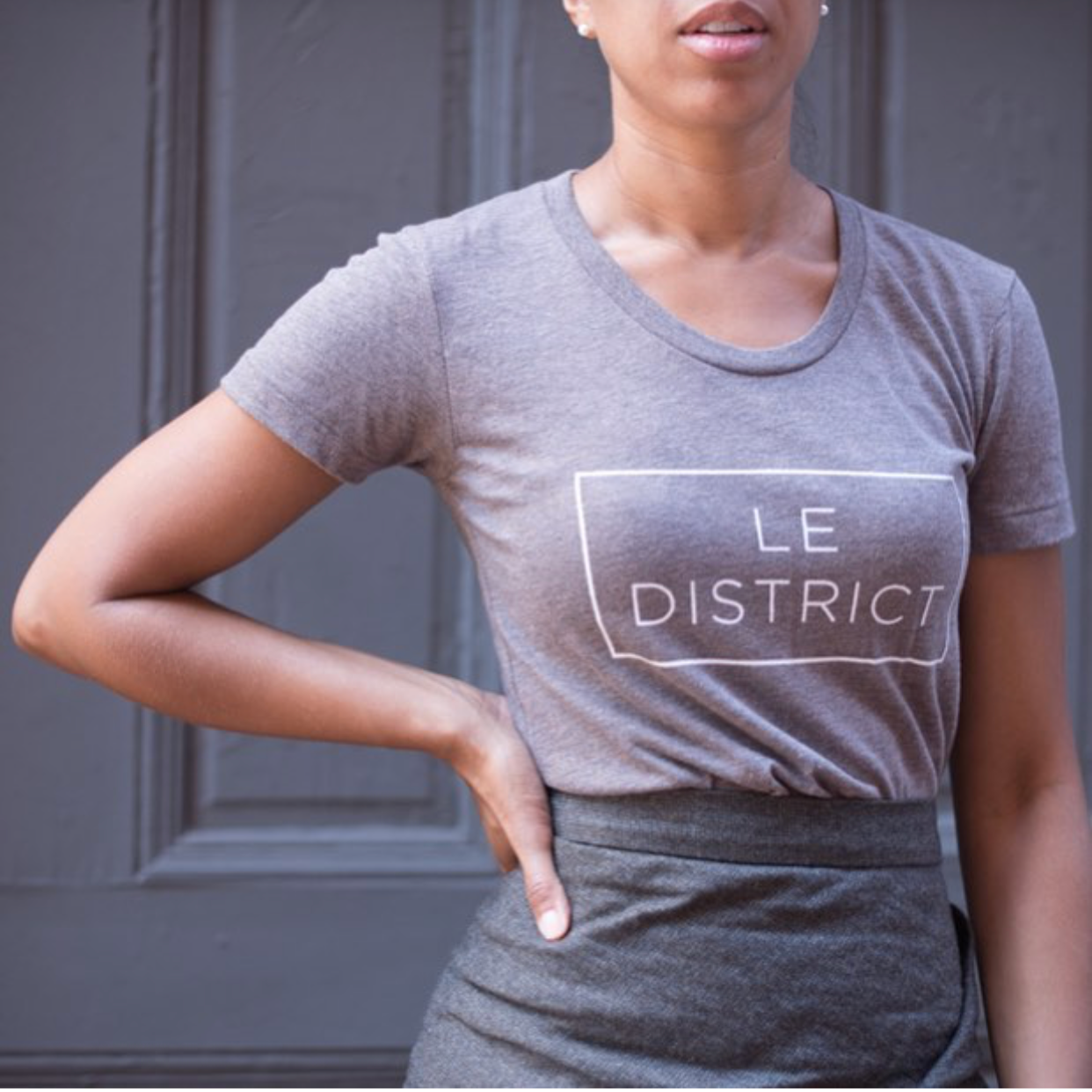 Close-up of a woman wearing a gray T-shirt with the words 'Le District' printed in white inside a rectangle, standing in front of a dark gray door.