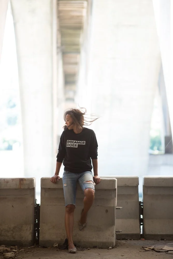 A woman in a black sweatshirt and ripped denim shorts sitting on a concrete barrier under a bridge or overpass.